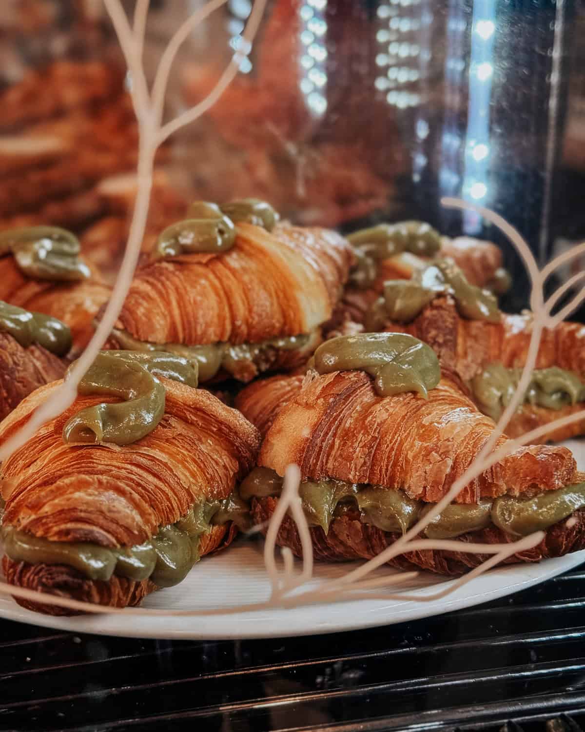 Close-up of a bakery display case showing a variety of pastries including croissants, cinnamon rolls, and sandwiches with small blackboards listing their names and prices.