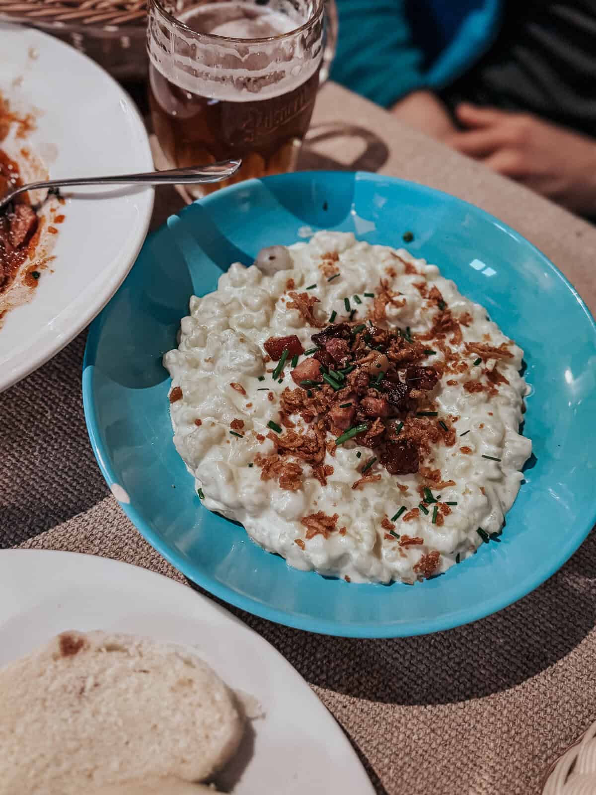 A bowl of cottage cheese noodles topped with crispy bacon bits and chopped chives, accompanied by a mug of beer and a partially visible plate of food in the background.