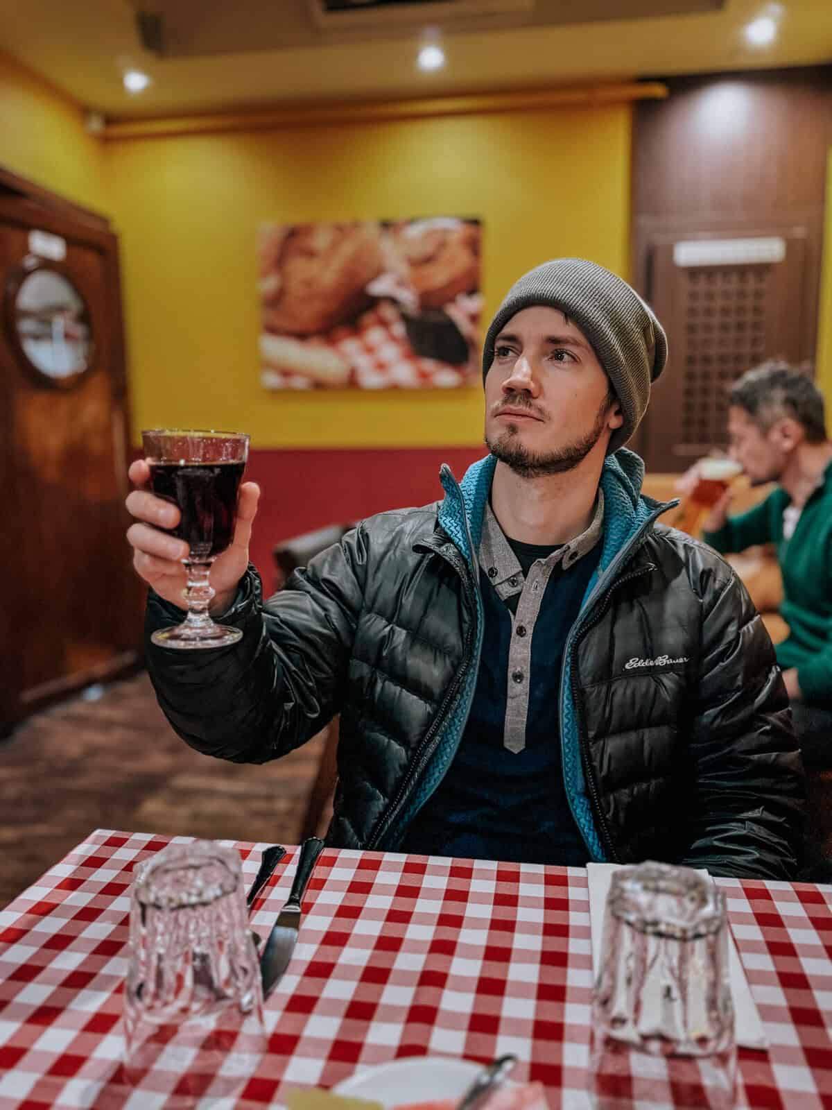 A person in a beanie and jacket raising a glass of dark red drink in a restaurant with yellow walls and red and white checkered tablecloths.