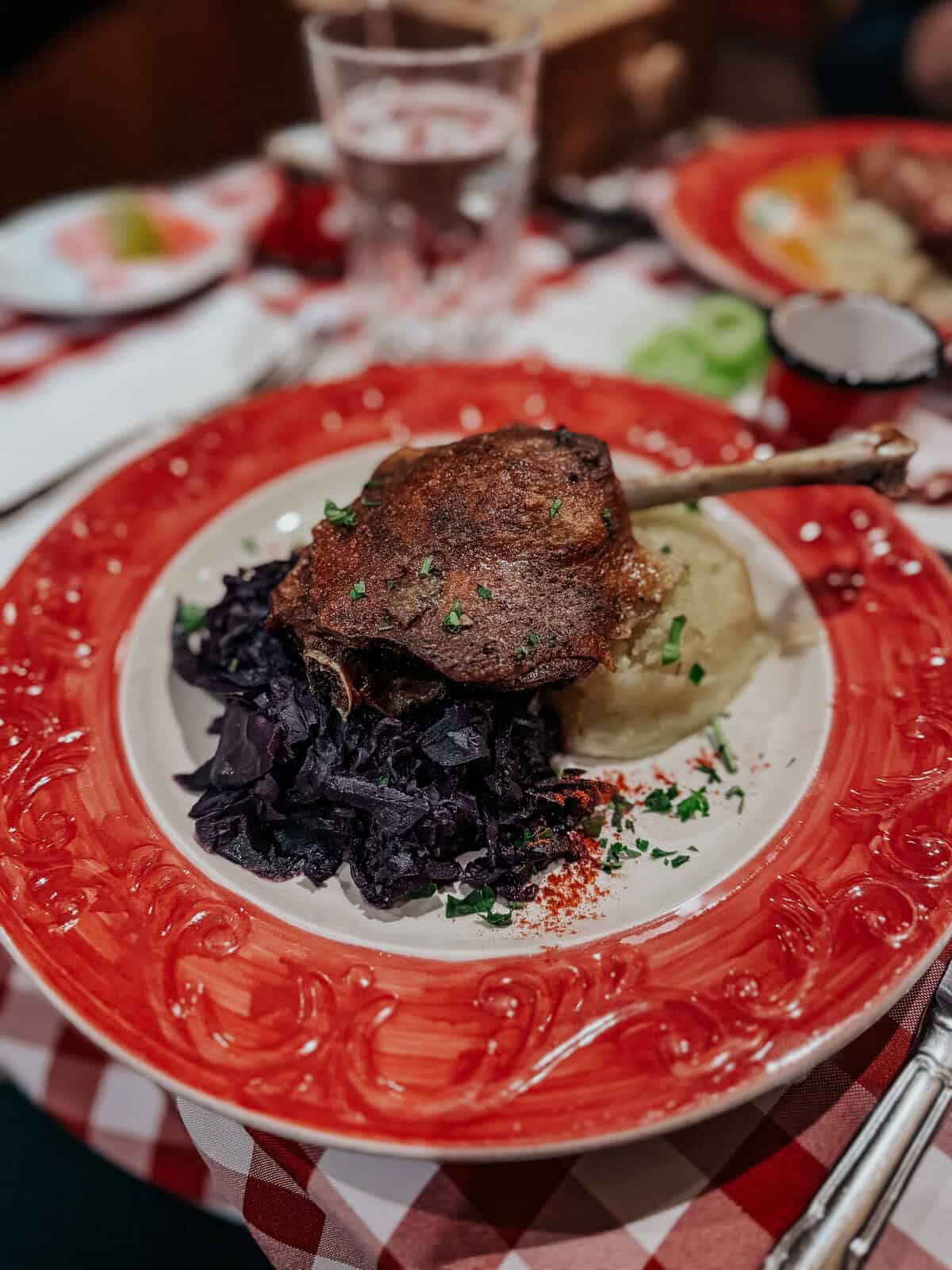 A table at a restaurant with a dish featuring roasted meat, purple cabbage, and mashed potatoes, all served on a decorative red and white plate.