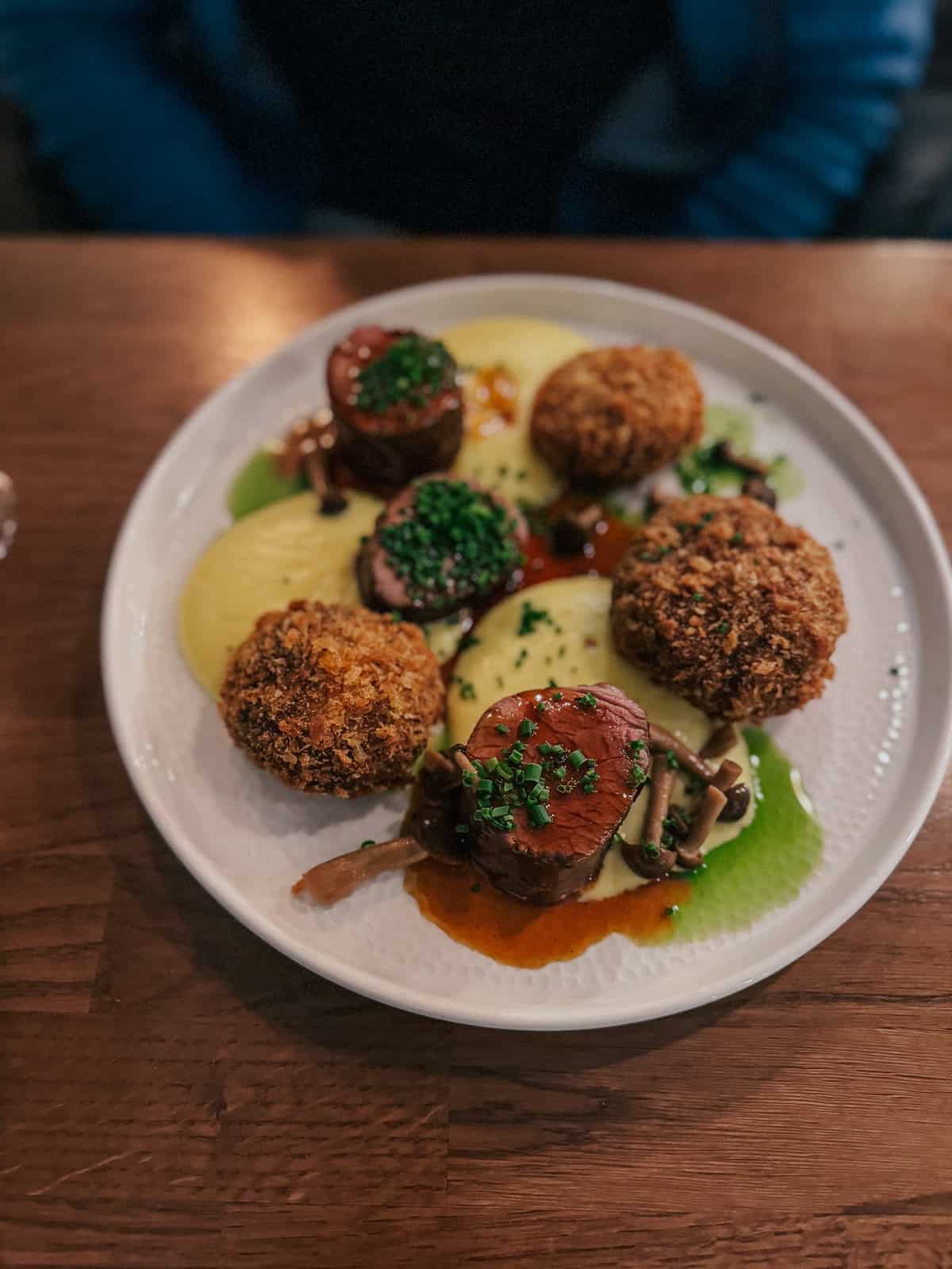 A plate of Mangalica pork medallions served with mashed potatoes, crispy croquettes, and garnished with fresh herbs.