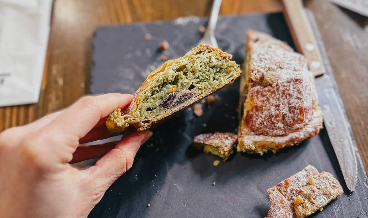 A hand holding a partially eaten croissant with a green filling and a hint of chocolate, placed on a dark plate.