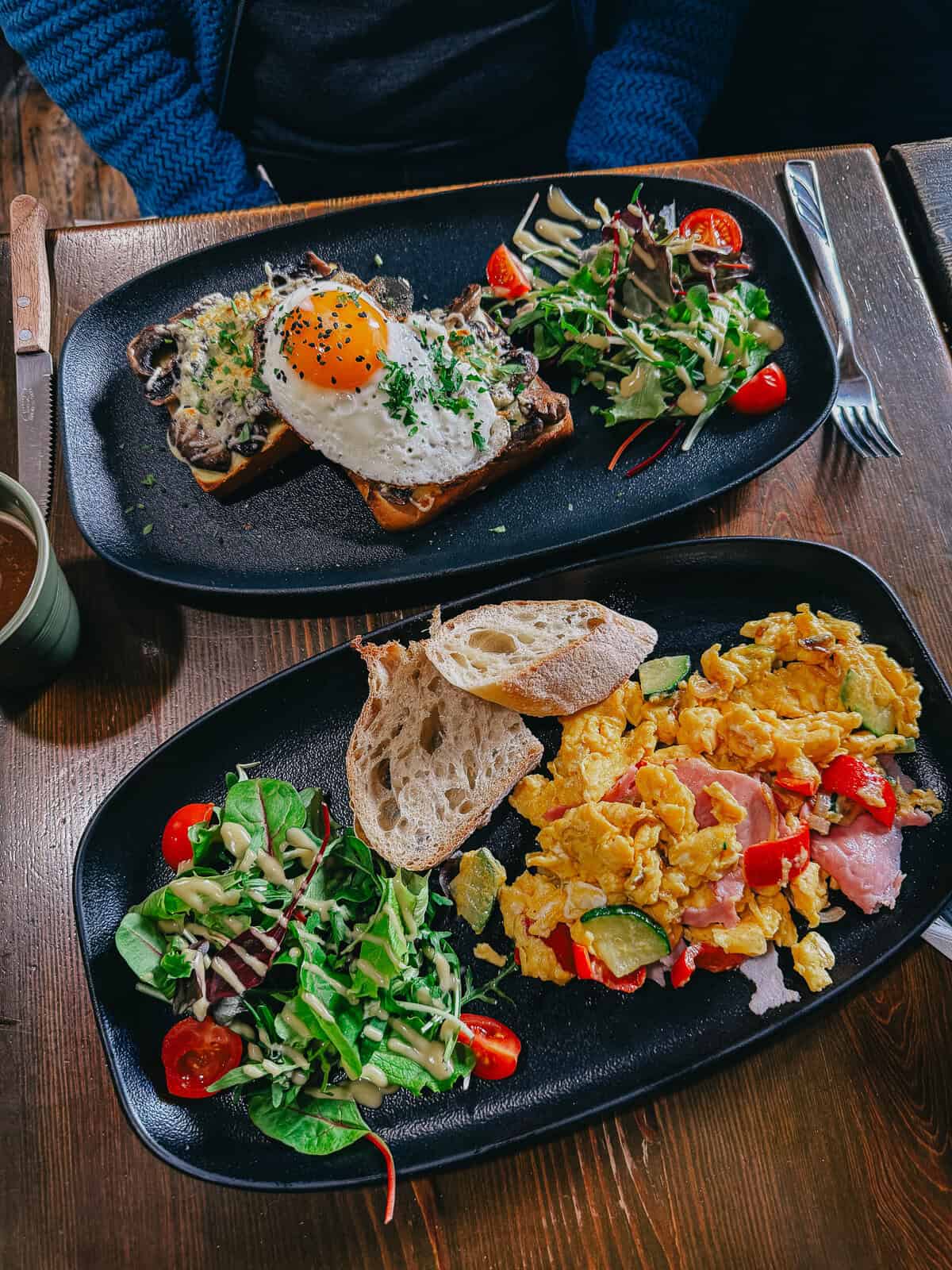 Two plates of breakfast food; one with scrambled eggs, salad, and bread, the other with toast topped with a fried egg, mushrooms, and salad.