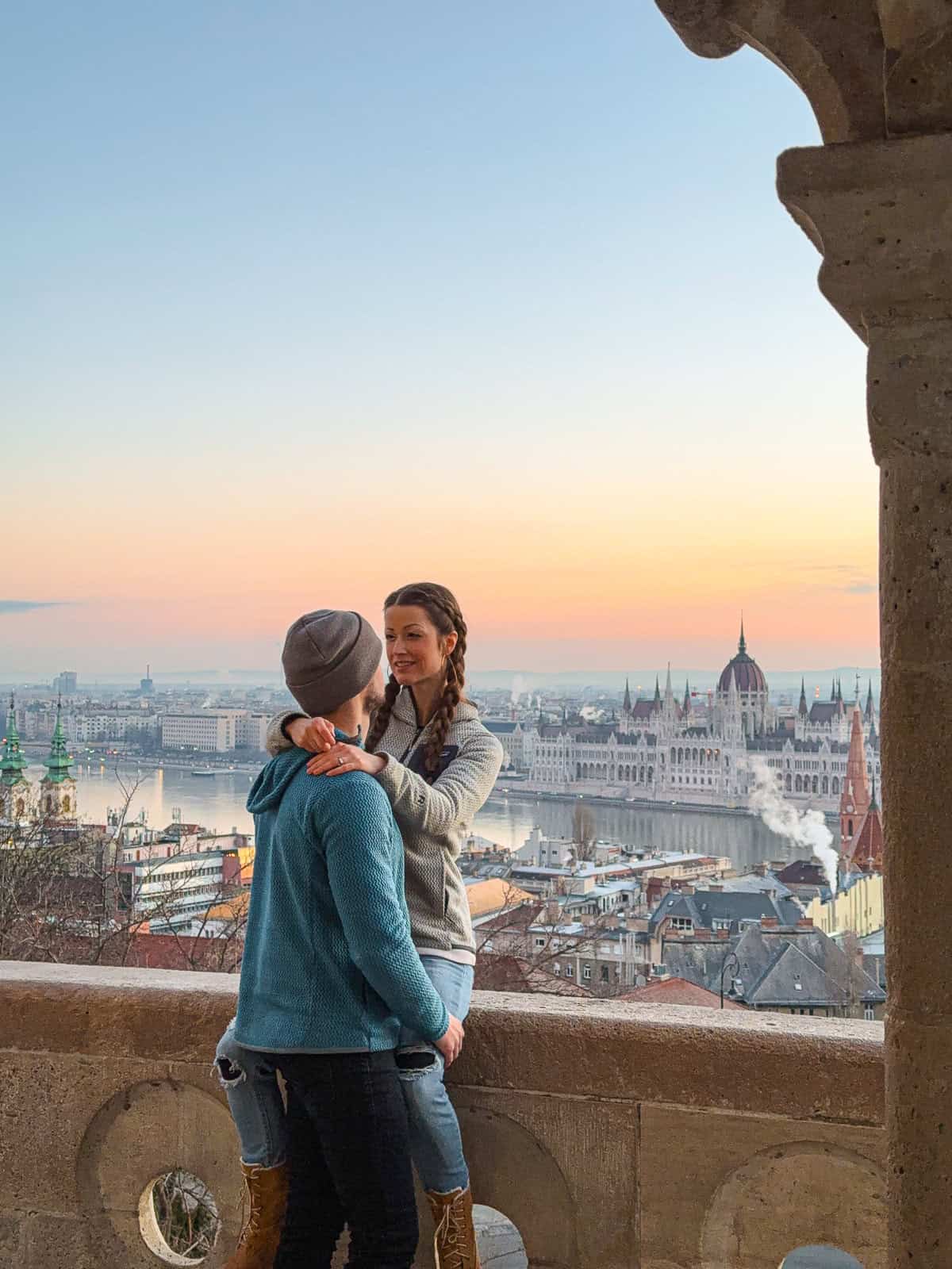 A couple embracing with a stunning panoramic view of Budapest at sunrise in the background, featuring the iconic Hungarian Parliament Building and the Danube River.