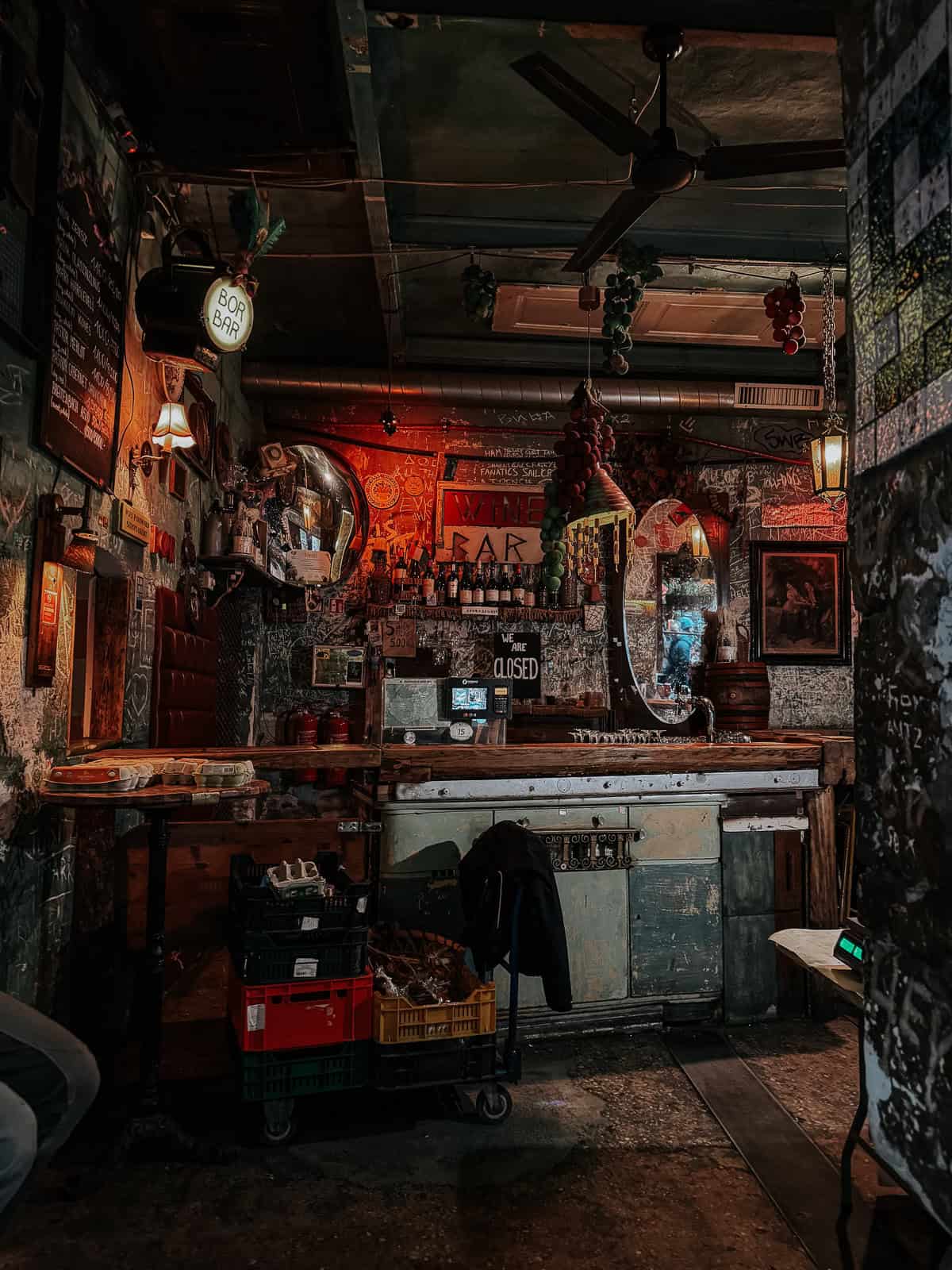 A dimly lit, rustic bar with a cozy atmosphere, featuring a vintage bar counter, hanging grapes decor, and a sign that reads 