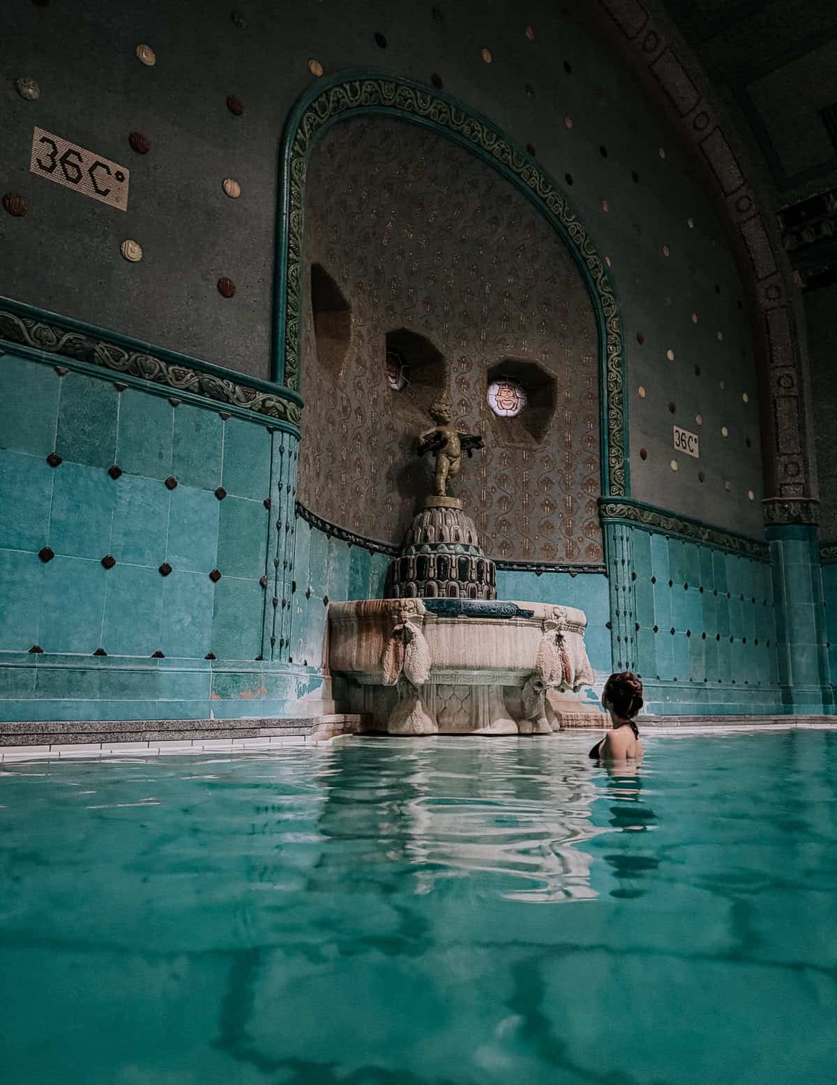 A person enjoys a thermal bath in a historic indoor pool, with a statue and 36°C temperature sign in the background, surrounded by teal tiles and ornate walls.