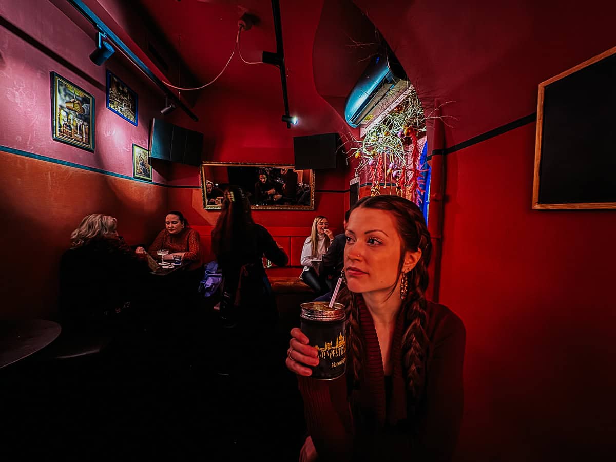 A woman with braided hair is sitting in a dimly lit bar with red walls, holding a drink and looking thoughtfully to the side. In the background, several other patrons are engaged in conversation at a table.