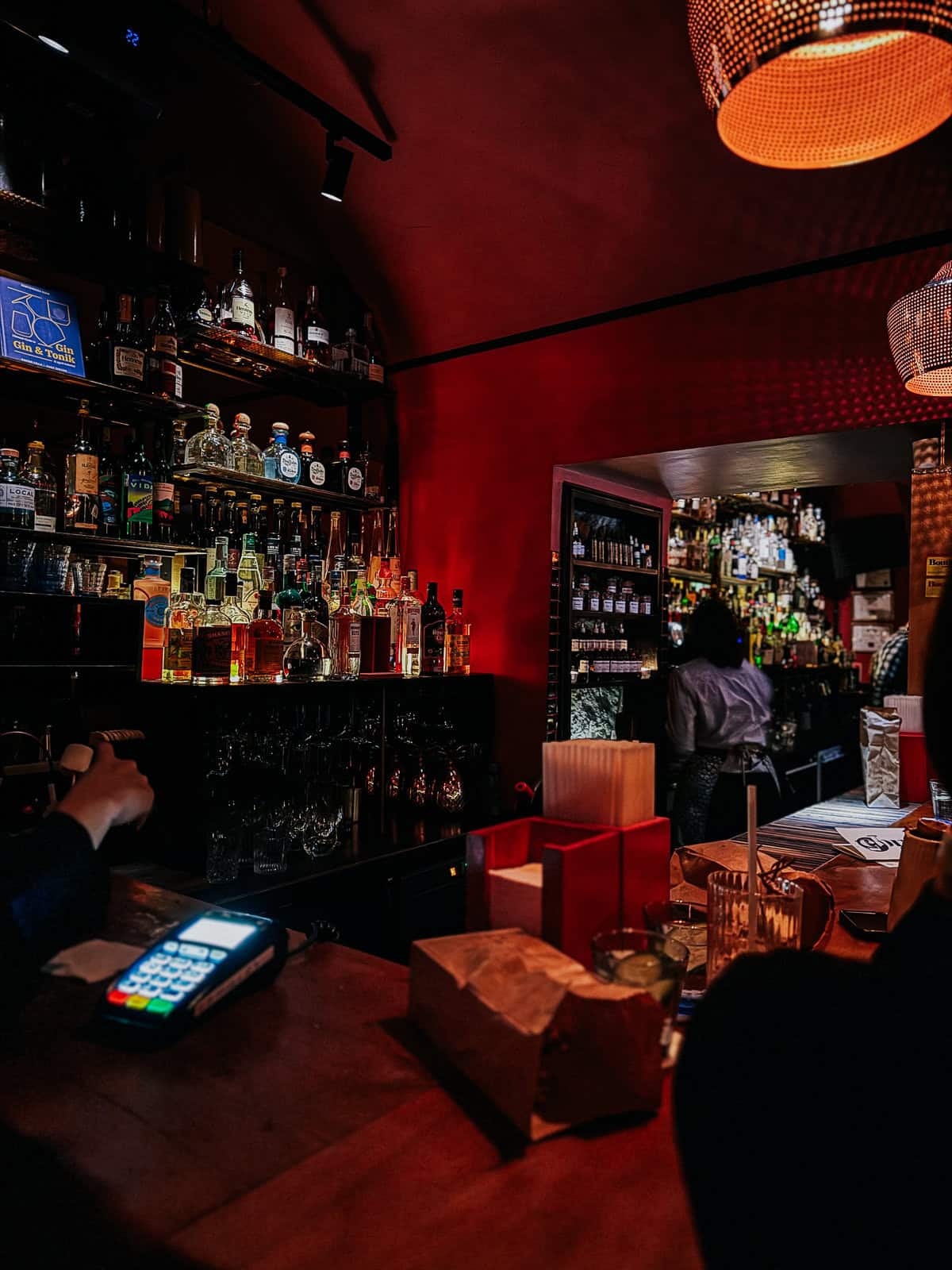 A well-stocked bar with various bottles of liquor displayed on shelves. A bartender is partially visible, working behind the bar, while the bar counter has a card payment terminal and some brown paper bags.