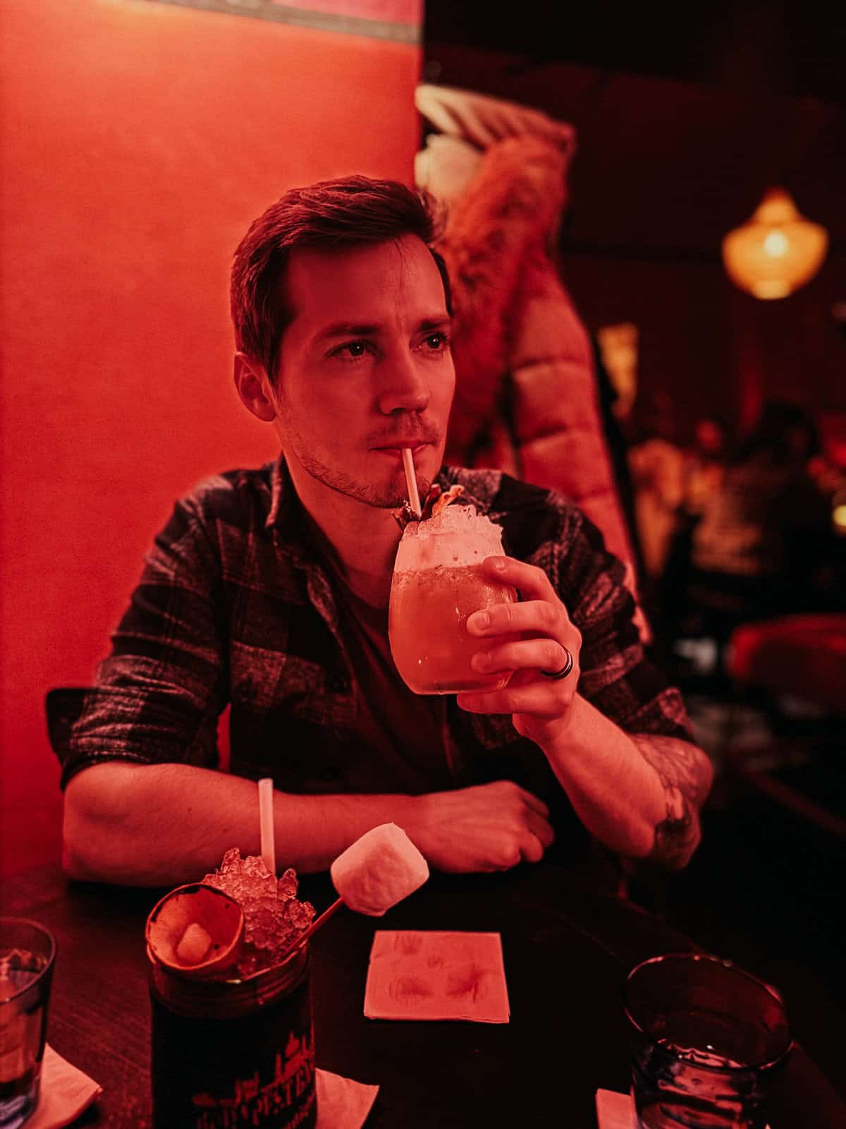 A man with a pensive expression is sipping a cocktail with a straw. The drink has an orange garnish and a large ice cube. The background is dimly lit, with warm red tones and a cozy atmosphere. A man with a pensive expression is sipping a cocktail with a straw. The drink has an orange garnish and a large ice cube. The background is dimly lit, with warm red tones and a cozy atmosphere