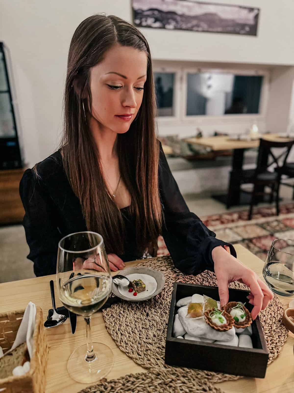 A woman with long brown hair is reaching into a black box of appetizers while a glass of white wine and a small dish with food are in front of her.