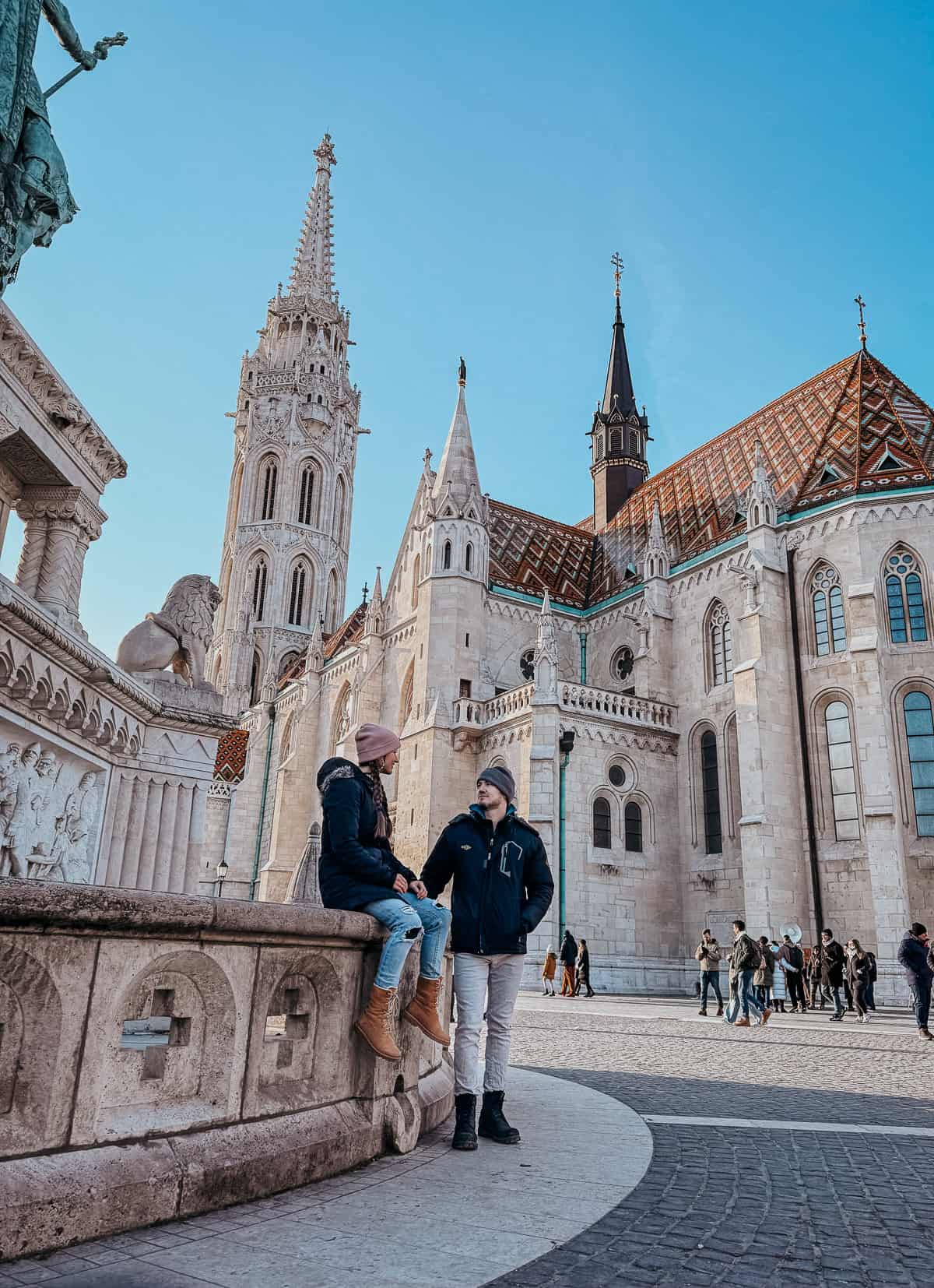 A couple dressed in winter clothing is sitting and standing on a ledge outside Matthias Church in Budapest, with its distinctive spires and roof tiles visible.