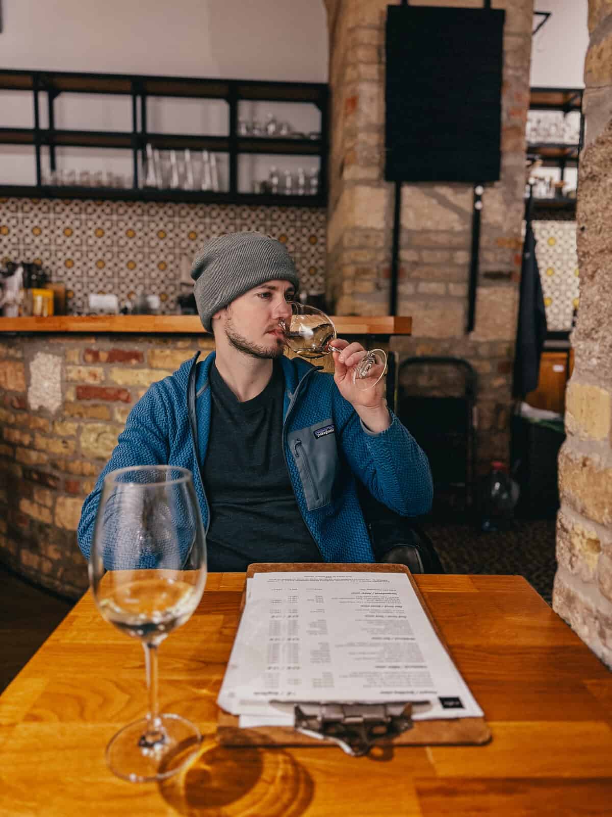 A man in a gray beanie and blue jacket is tasting white wine inside a rustic restaurant with brick and tiled walls.