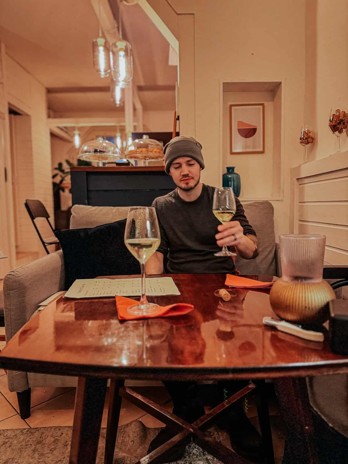 A man in a gray beanie is sitting at a table in a cozy restaurant, holding a glass of white wine. The table has a menu and another glass of wine.