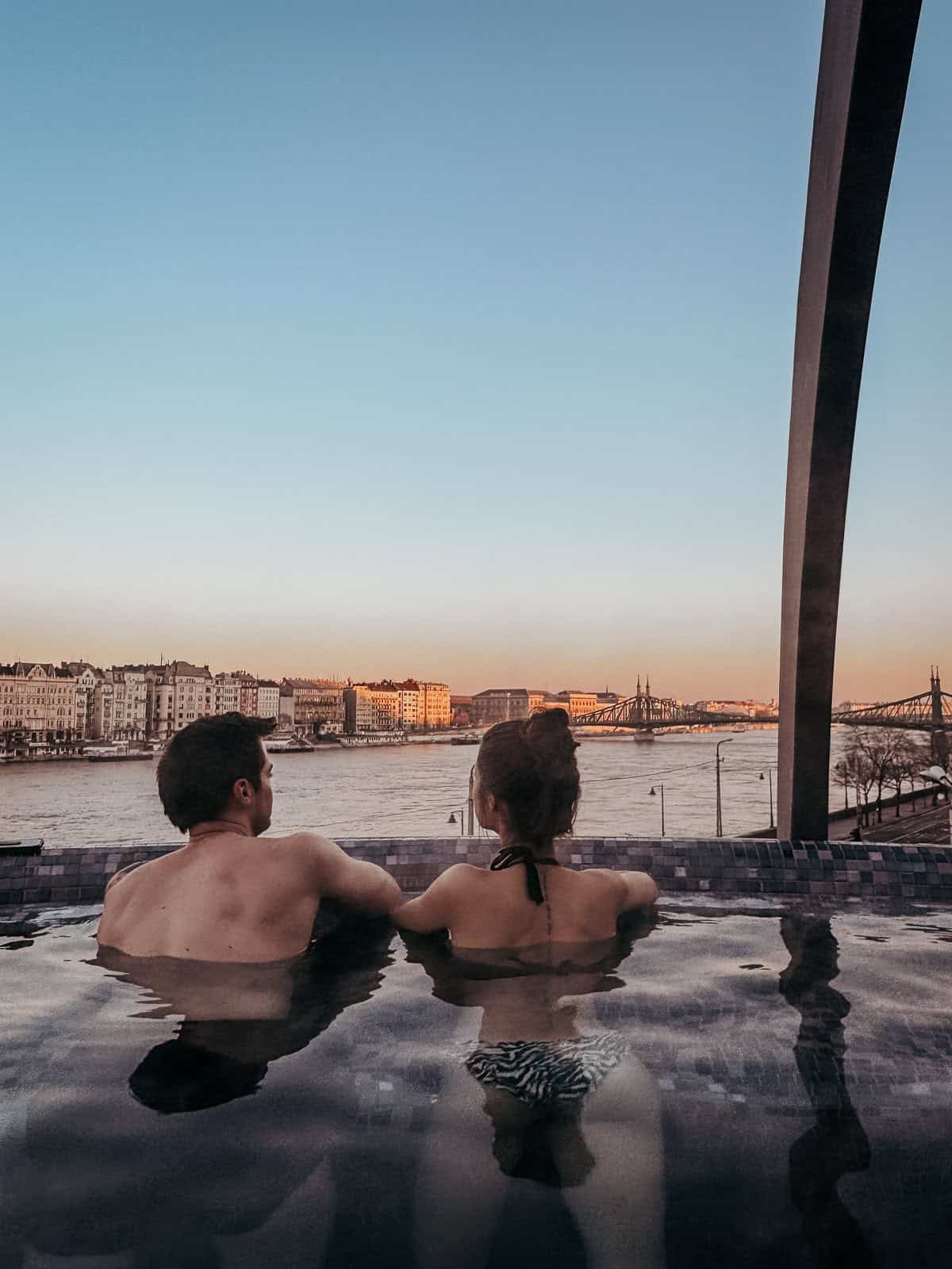 A couple is relaxing in an outdoor hot tub overlooking the Danube River and Budapest at sunset.