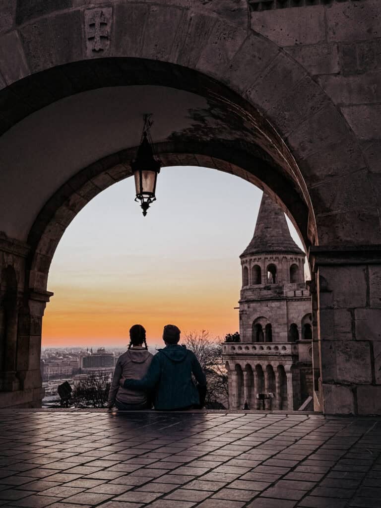 A couple is sitting and enjoying the sunset under an archway at Fisherman's Bastion in Budapest, with the city skyline in the background.
