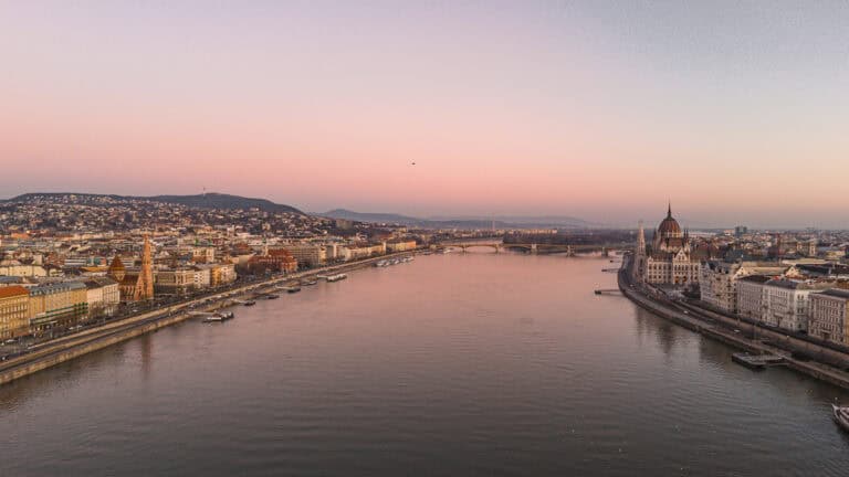 An aerial view of Budapest at sunrise, showcasing the Ferris wheel, historic buildings, and the Danube River, with the hills in the distance.