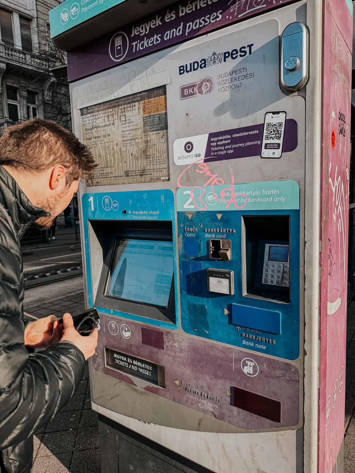A man using a ticket machine in Budapest, with signs indicating payment options, including bank cards and coins.