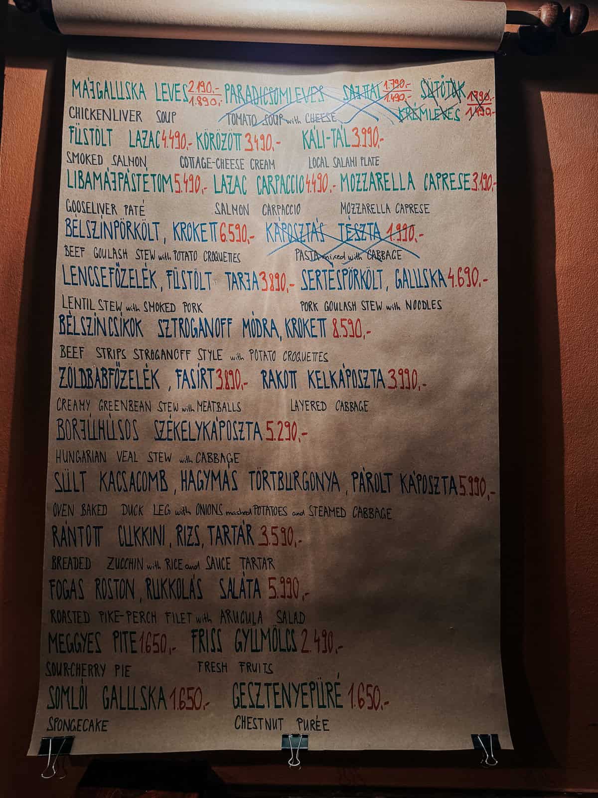 A handwritten menu on a roll of brown paper in a restaurant, listing various dishes in Hungarian with prices, including chicken liver soup, smoked salmon, goose liver pâté, beef goulash, and chestnut purée.