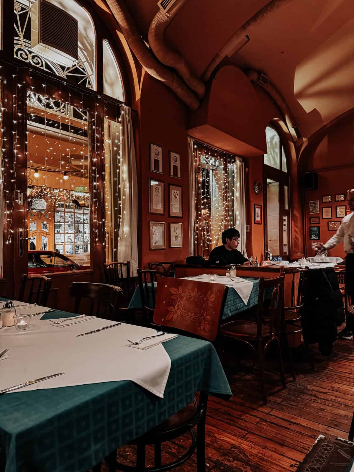 The interior of a cozy restaurant with teal tablecloths, white table runners, wooden chairs, and twinkling string lights hanging around the windows. A man is sitting alone at a table, and the room is decorated with framed awards and certificates.