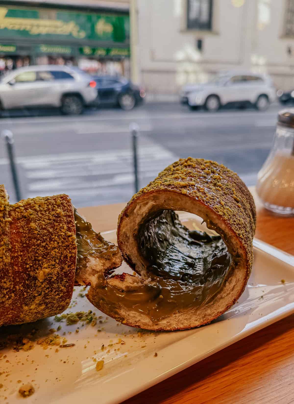 A close-up of a chimney cake covered in ground nuts, split open to reveal a filling of greenish cream, with a street view visible through a cafe window.