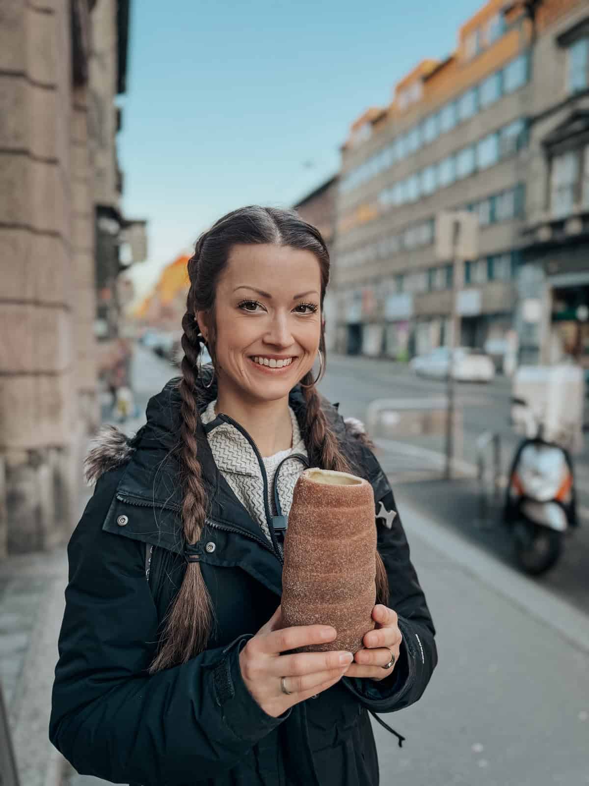 A smiling woman holding a chimney cake, standing on a city street with buildings and traffic in the background.