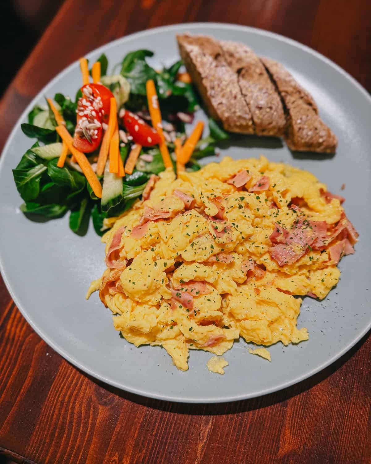 a blue plate piled high with scrambled eggs with ham, a small side salad and 2 pieces of baguette