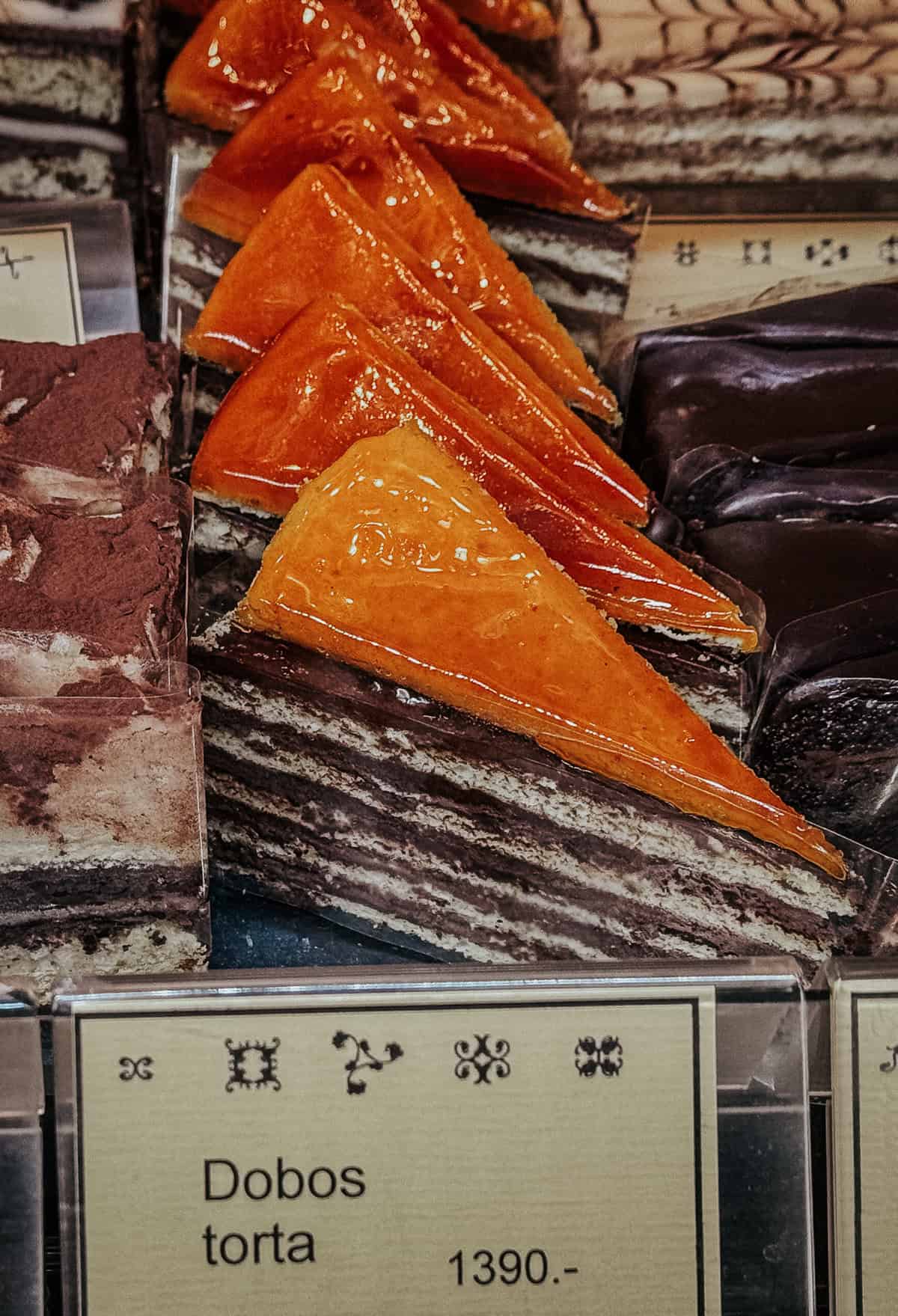 Slices of Dobos torta, a Hungarian layered cake topped with caramel glaze, displayed in a bakery with a price tag of 1390 HUF.