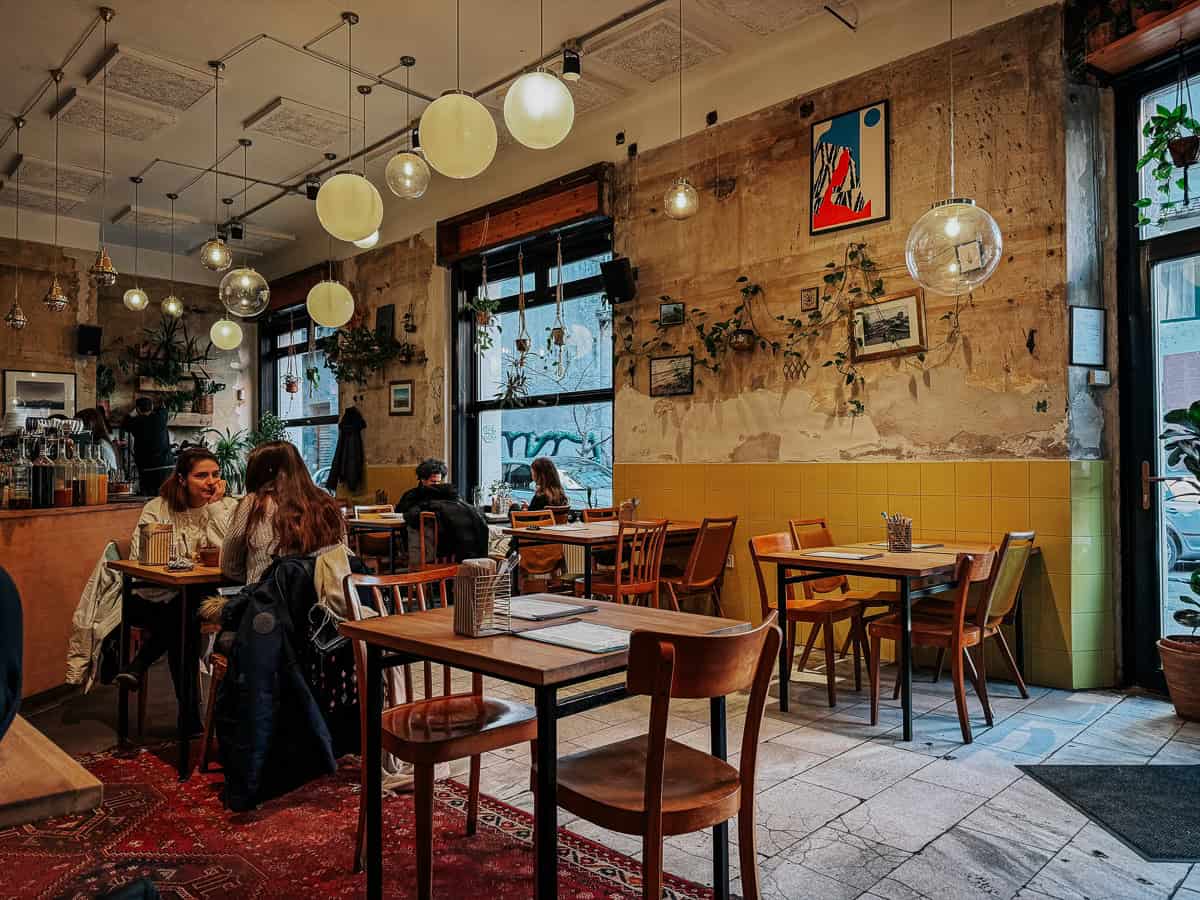 A cozy cafe interior with warm lighting, wooden furniture, and various plants and artwork adorning the walls, creating a welcoming atmosphere.