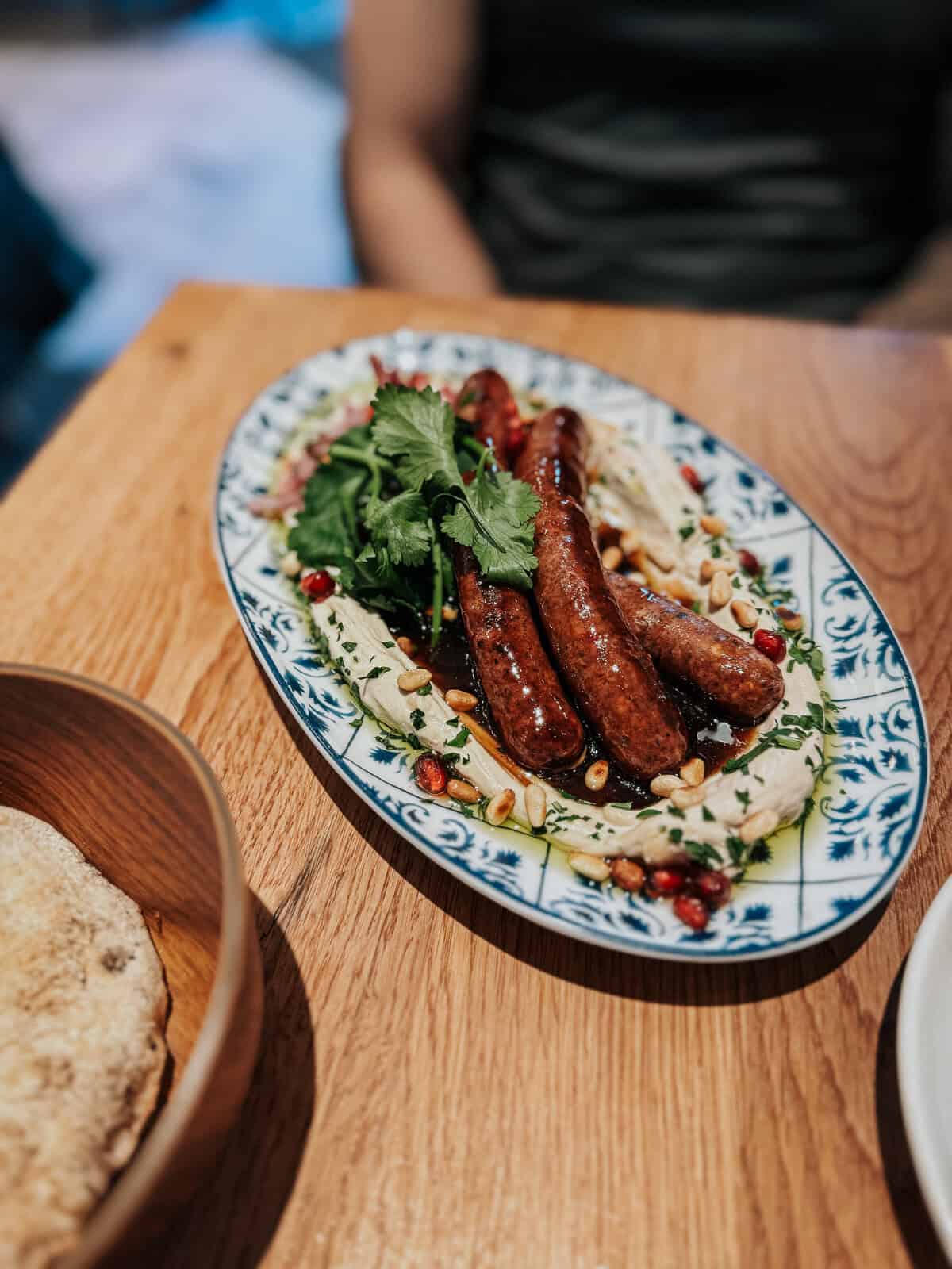 A platter of grilled sausages served on a bed of hummus, garnished with herbs and pomegranate seeds, with a side of flatbread.