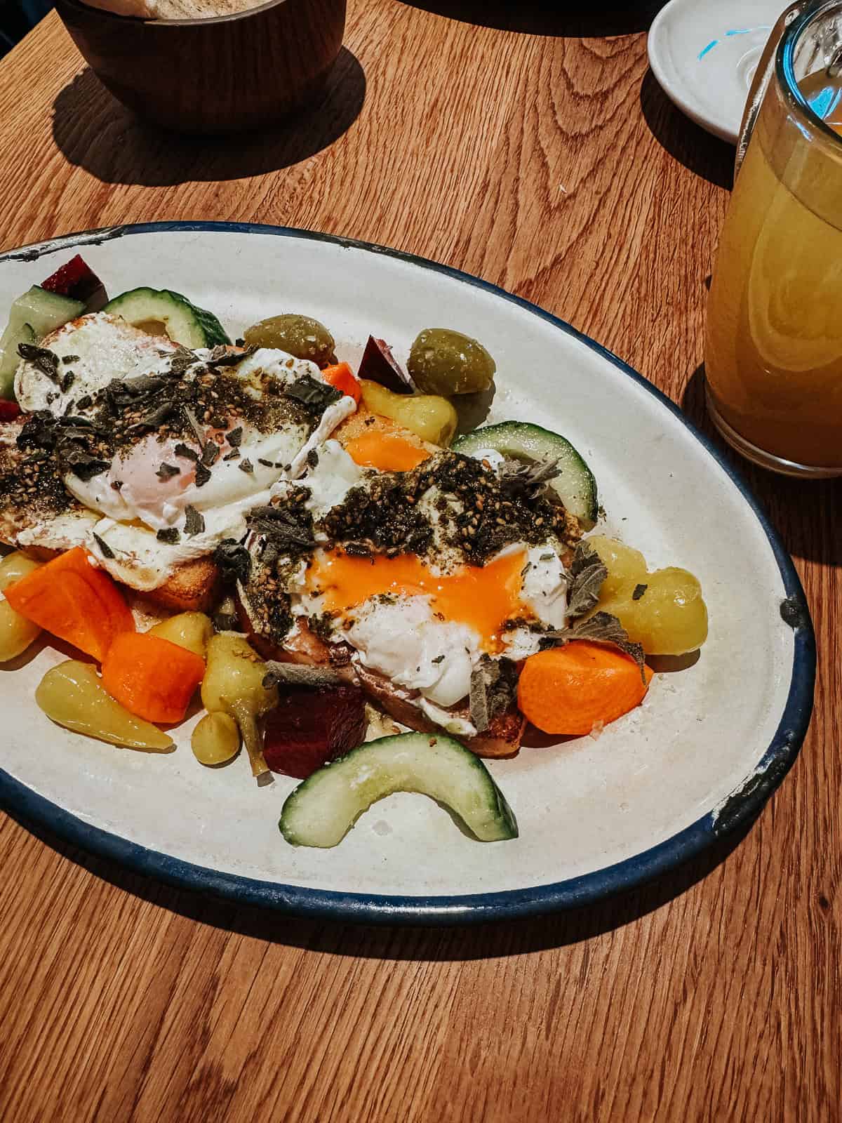 A colorful brunch plate with two eggs, diced vegetables, and a mix of pickles, set on a wooden table with a glass of juice.