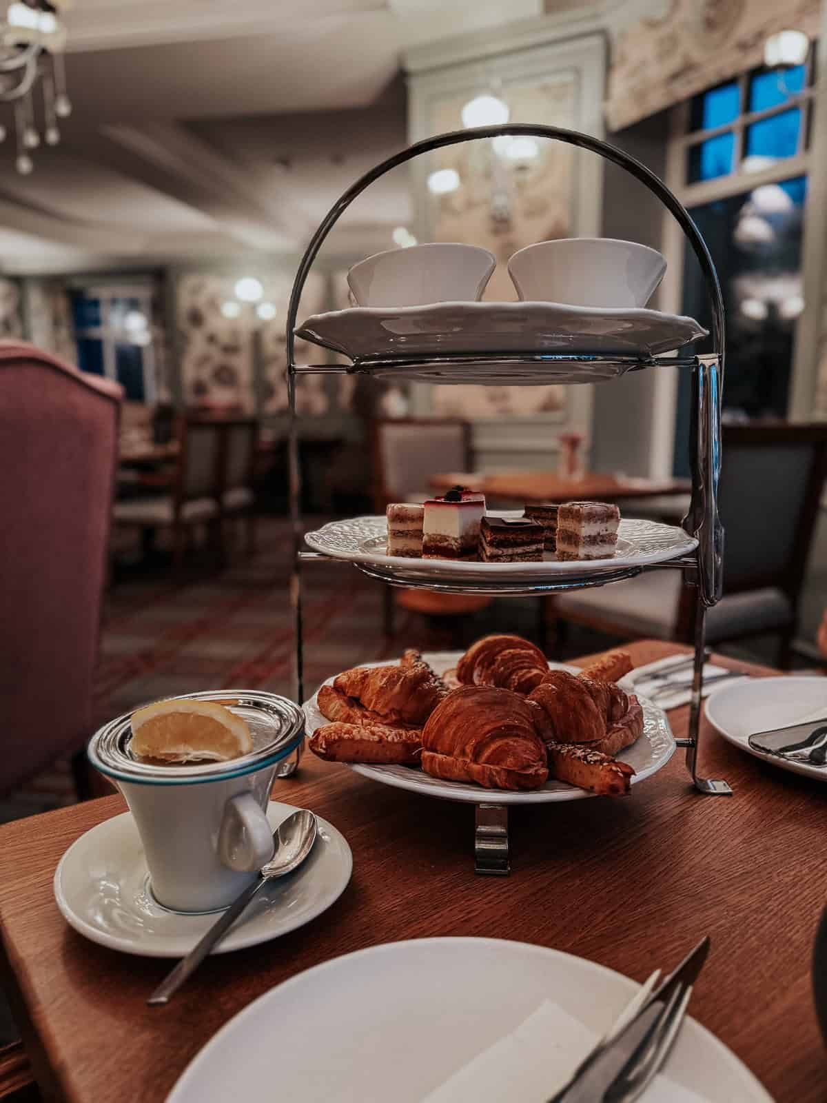 A tiered serving tray with a selection of pastries and desserts, alongside a cup of tea with a slice of lemon, set in an elegant dining room.