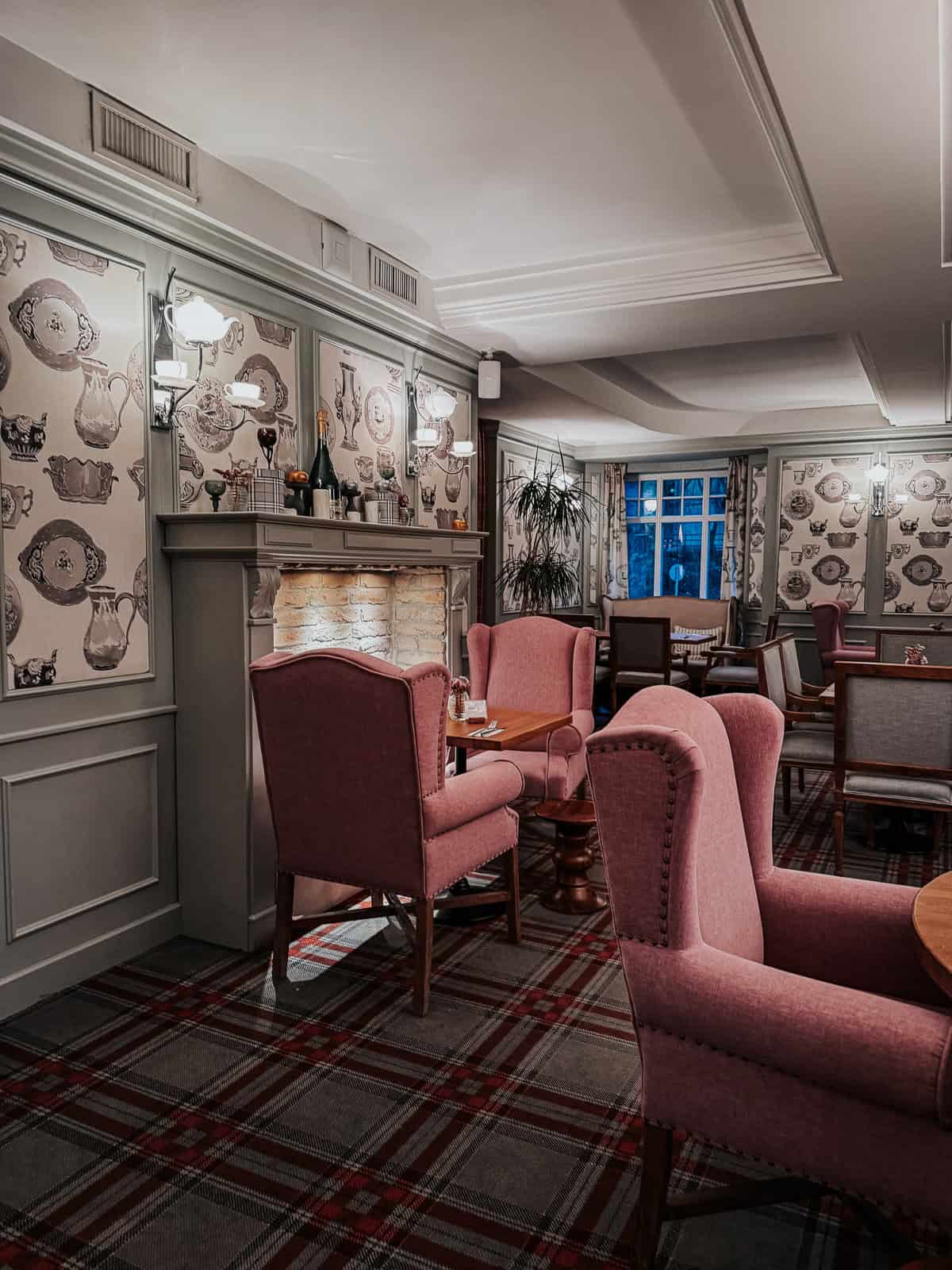 An elegant, cozy dining room with pink upholstered chairs, patterned wallpaper, and a stone fireplace, creating a warm and inviting atmosphere.