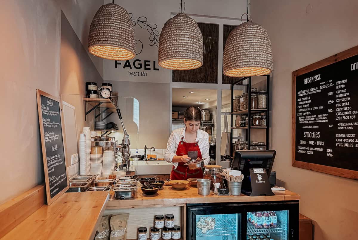 A barista behind the counter of Fagel café, preparing a dish under warm hanging lights, with shelves of ingredients and a menu board in the background.