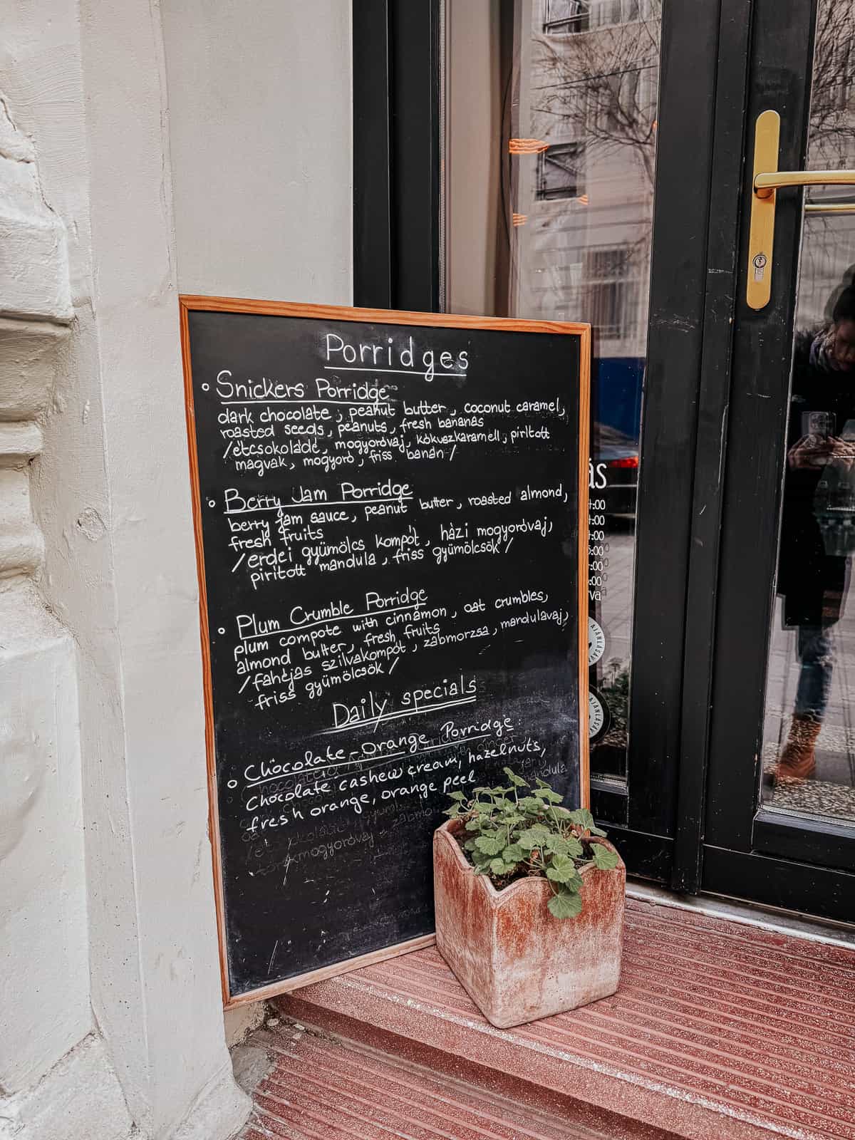 A chalkboard menu outside a café listing various porridge options and daily specials, next to a small potted plant.