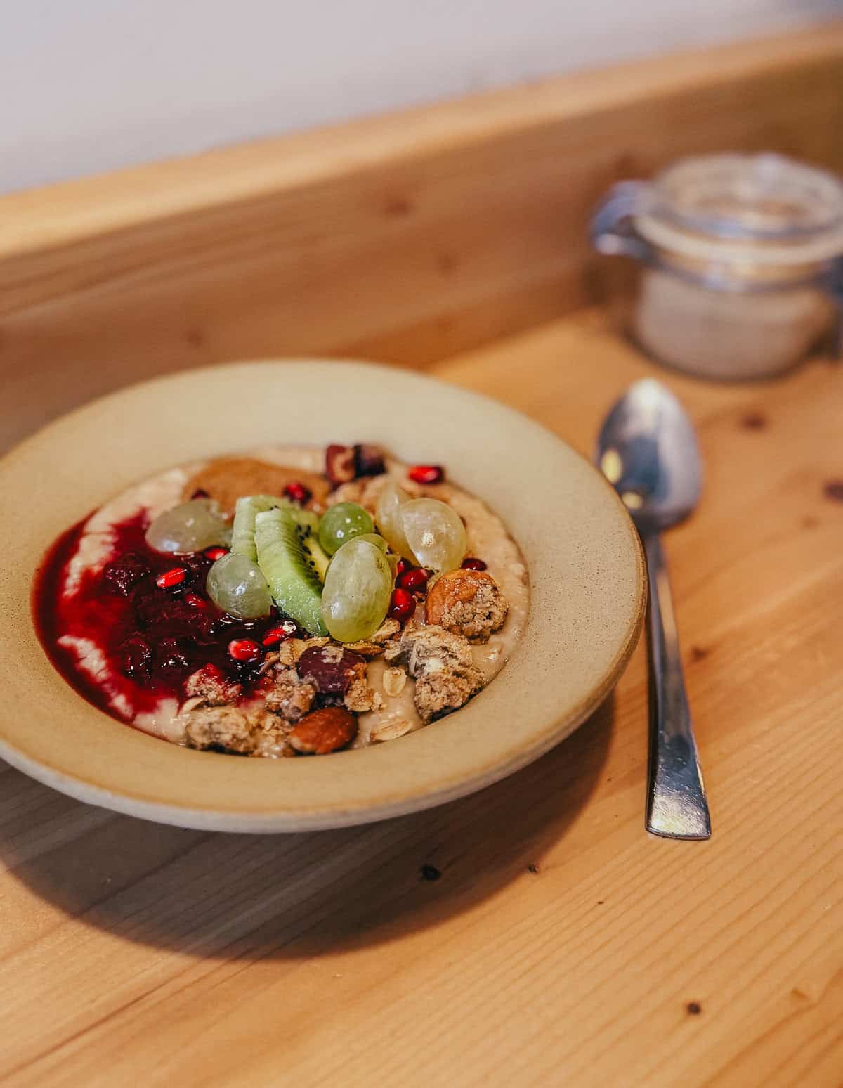 A bowl of oatmeal topped with kiwi slices, green grapes, pomegranate seeds, granola, and a berry compote, with a spoon beside it on a wooden table.