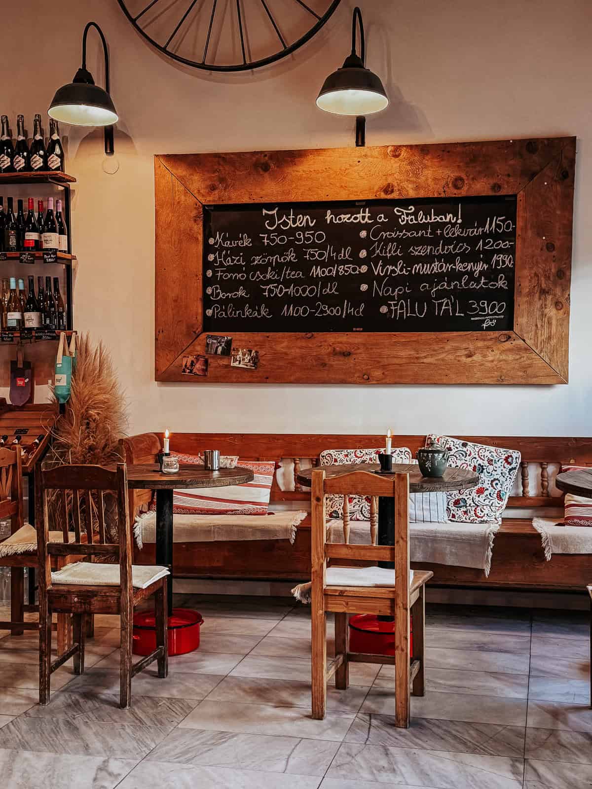 A rustic cafe interior with wooden tables and chairs, featuring a blackboard menu with various drink and food options in Hungarian.