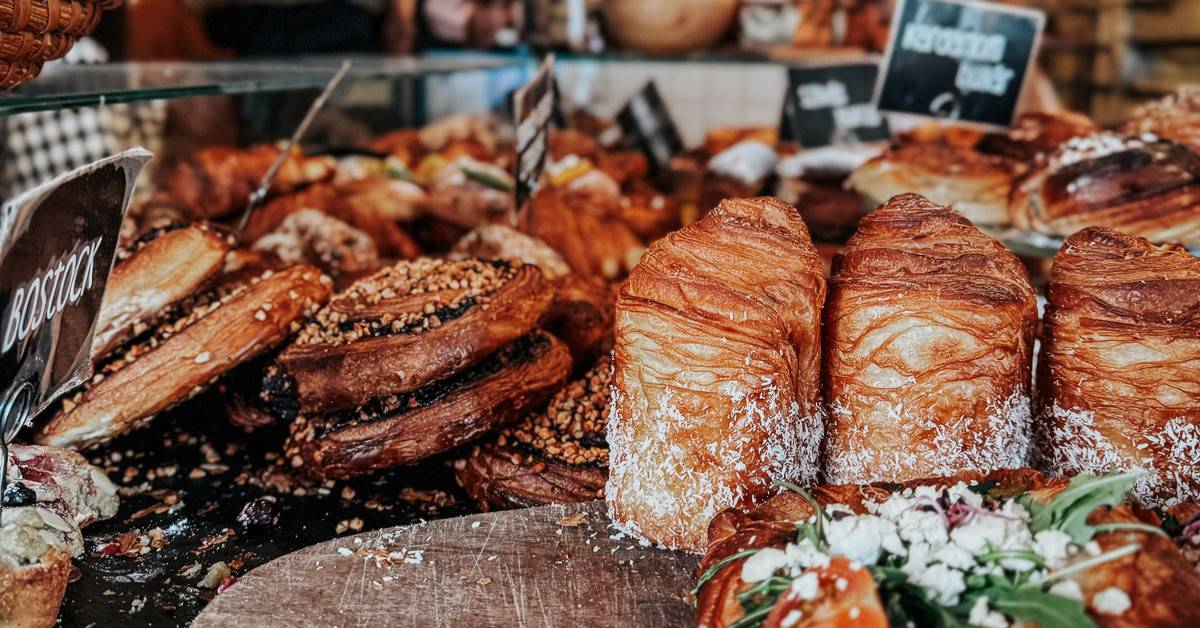 An assortment of pastries and baked goods is displayed on a wooden board inside a bakery, with various pastries labeled and customers in the background.