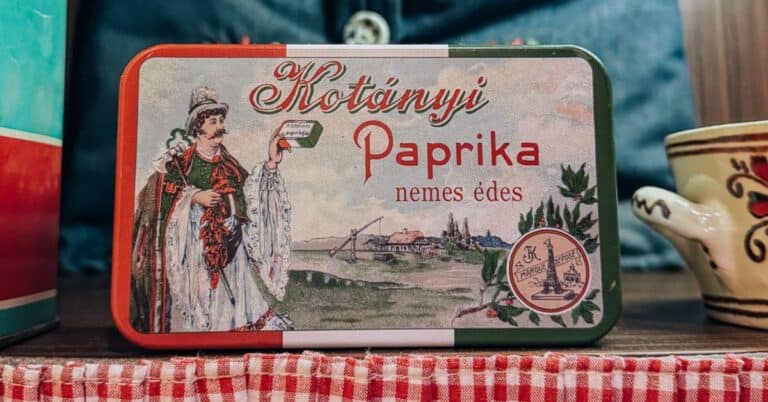 A vintage-style tin of Hungarian paprika, labeled "Kotányi Paprika," displayed on a shelf with red-and-white checkered fabric.