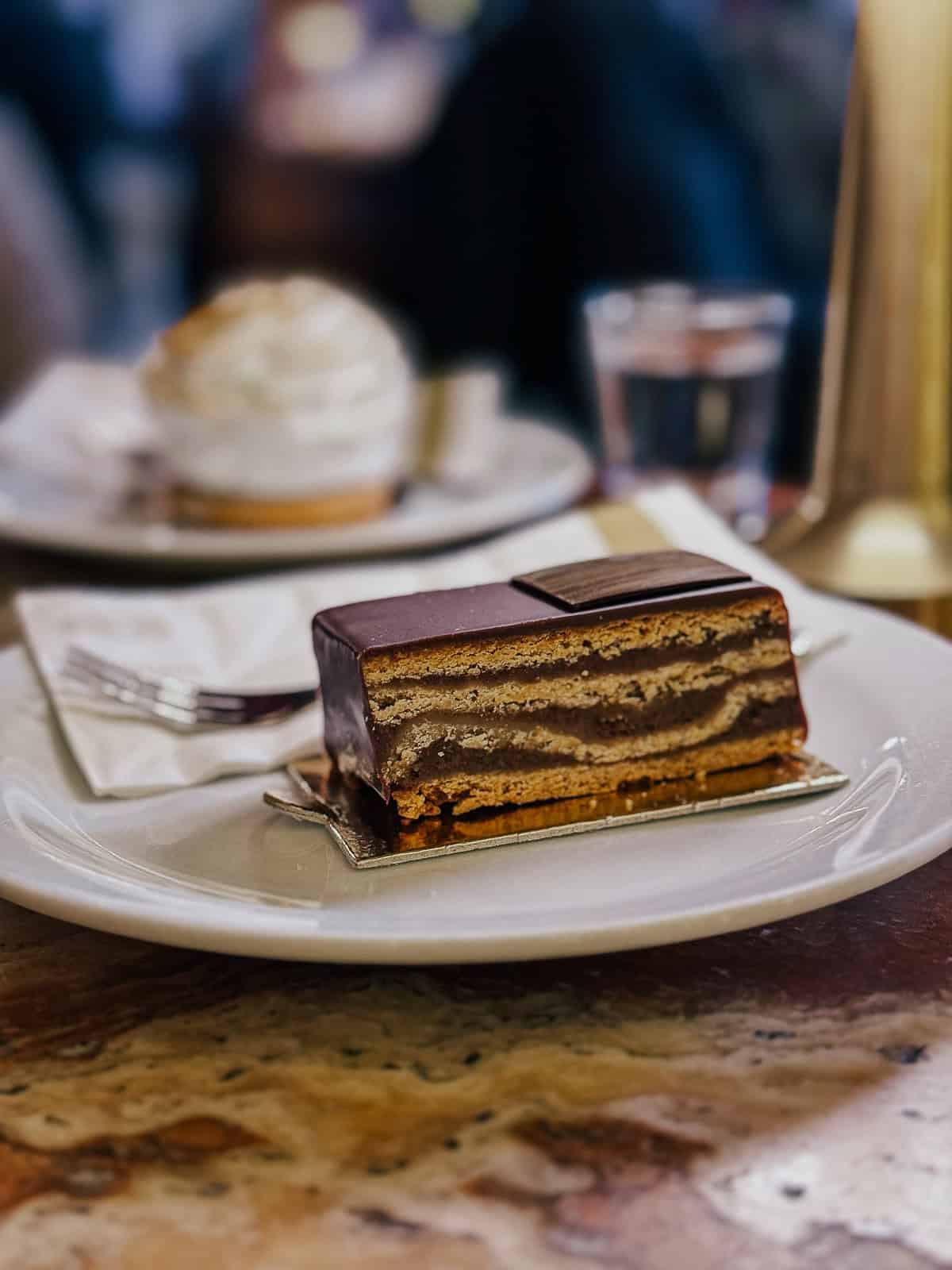 A slice of Gerbeaud cake, a layered Hungarian dessert with chocolate, walnuts, and apricot jam, served on a white plate in a cafe setting.