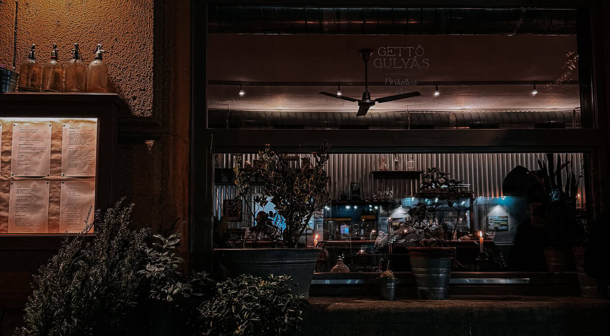 A dimly lit view of a restaurant window at night, with plants in the foreground and a warm, cozy interior visible inside.