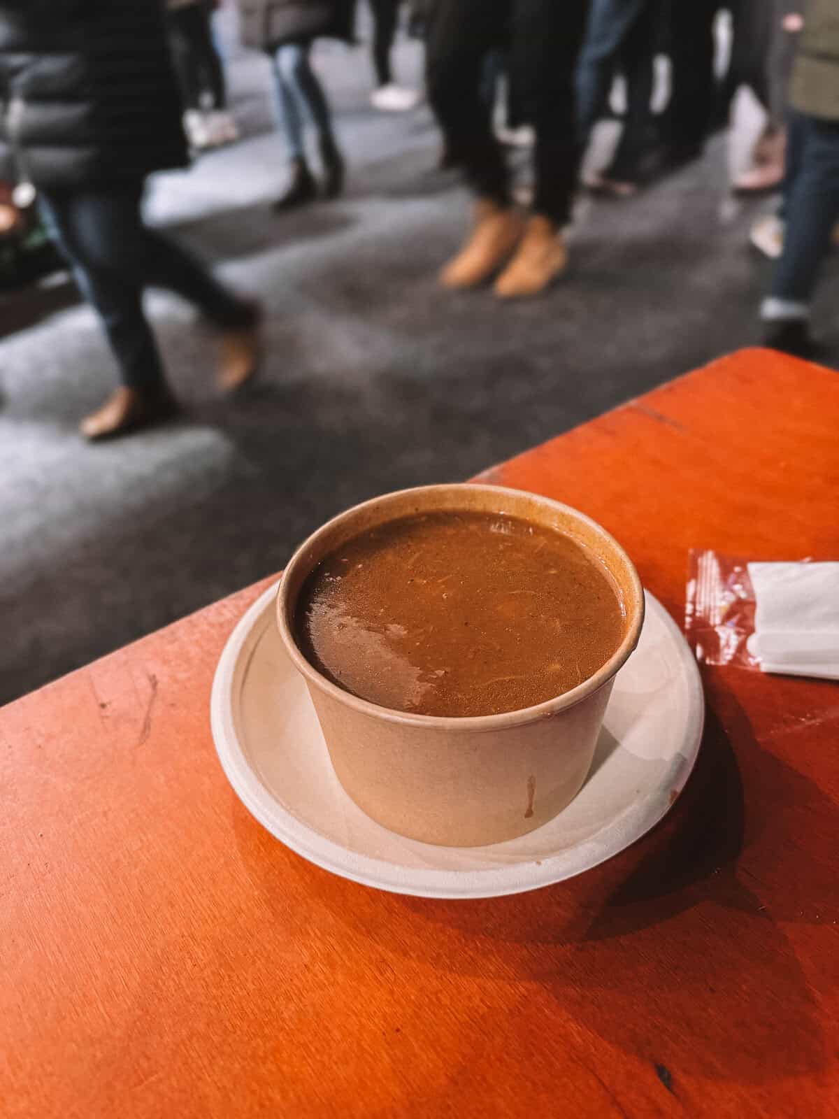 A bowl of goulash soup served in a paper cup on an orange table, with blurred people walking in the background at a market or festival.