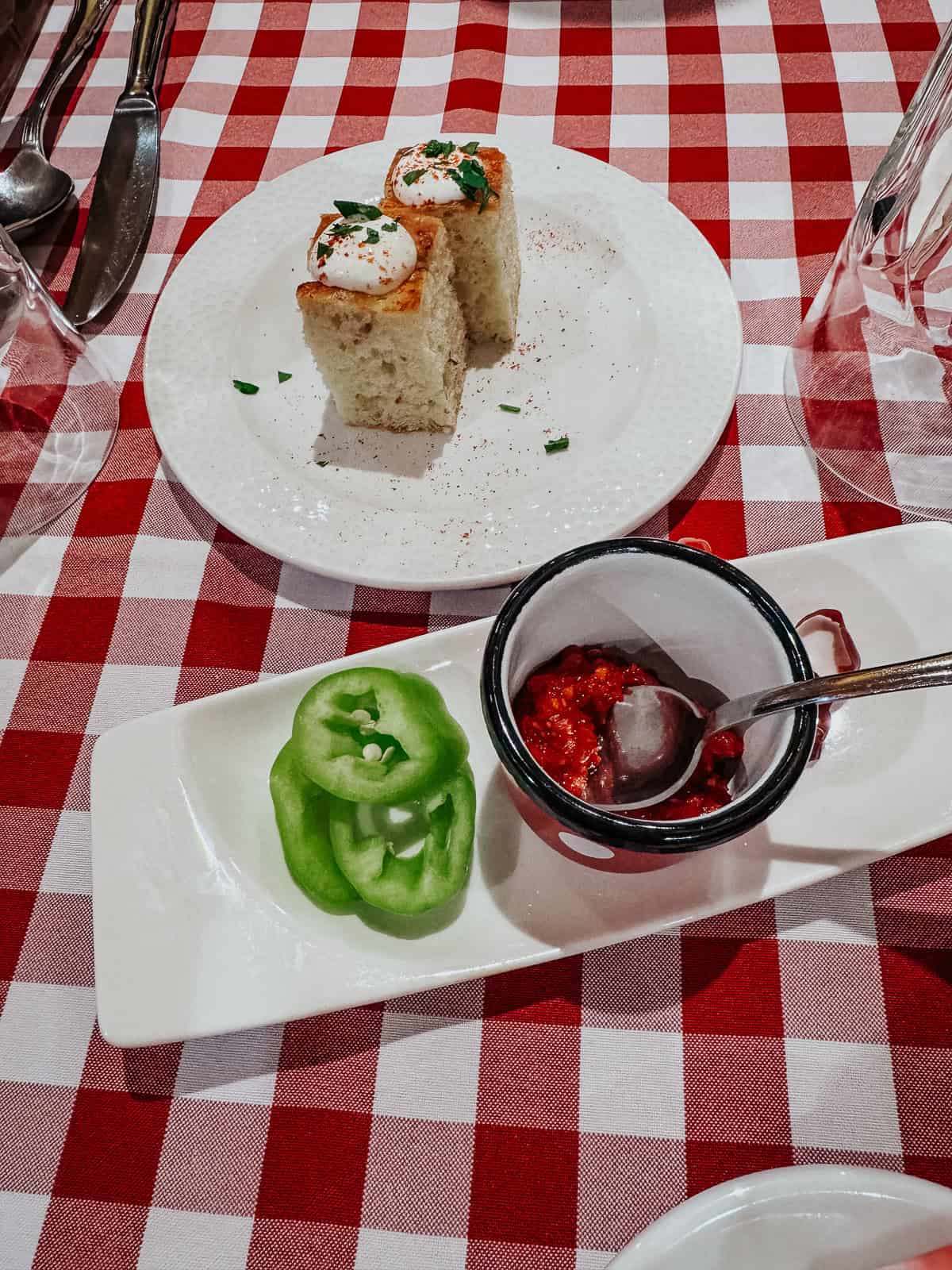 A table set with slices of bread topped with a dollop of sour cream, green pepper slices, and a small dish of red sauce, on a red-and-white checkered tablecloth.