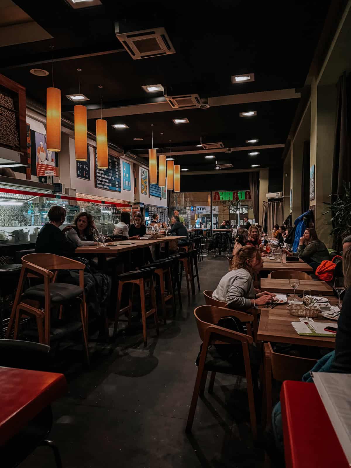 The interior of a wine bar with patrons sitting at high tables and chairs, illuminated by warm pendant lights. The bar features a modern design with a display of various wines and a lively atmosphere.