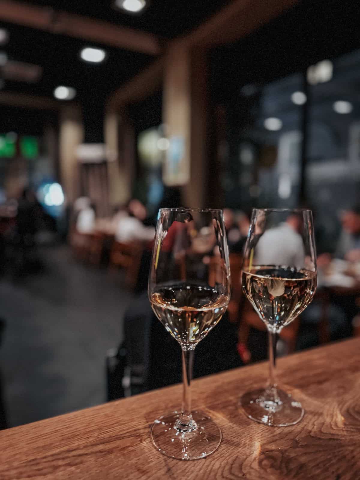 Two glasses of white wine on a wooden counter in a dimly lit restaurant, with blurred diners in the background creating a cozy and intimate ambiance.