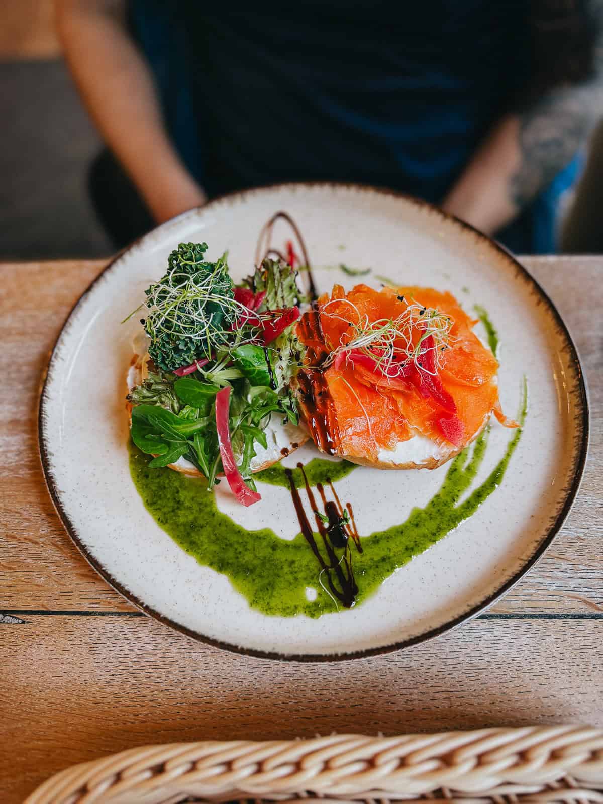 A plate of smoked salmon with greens and sprouts, drizzled with green sauce, on a wooden table.