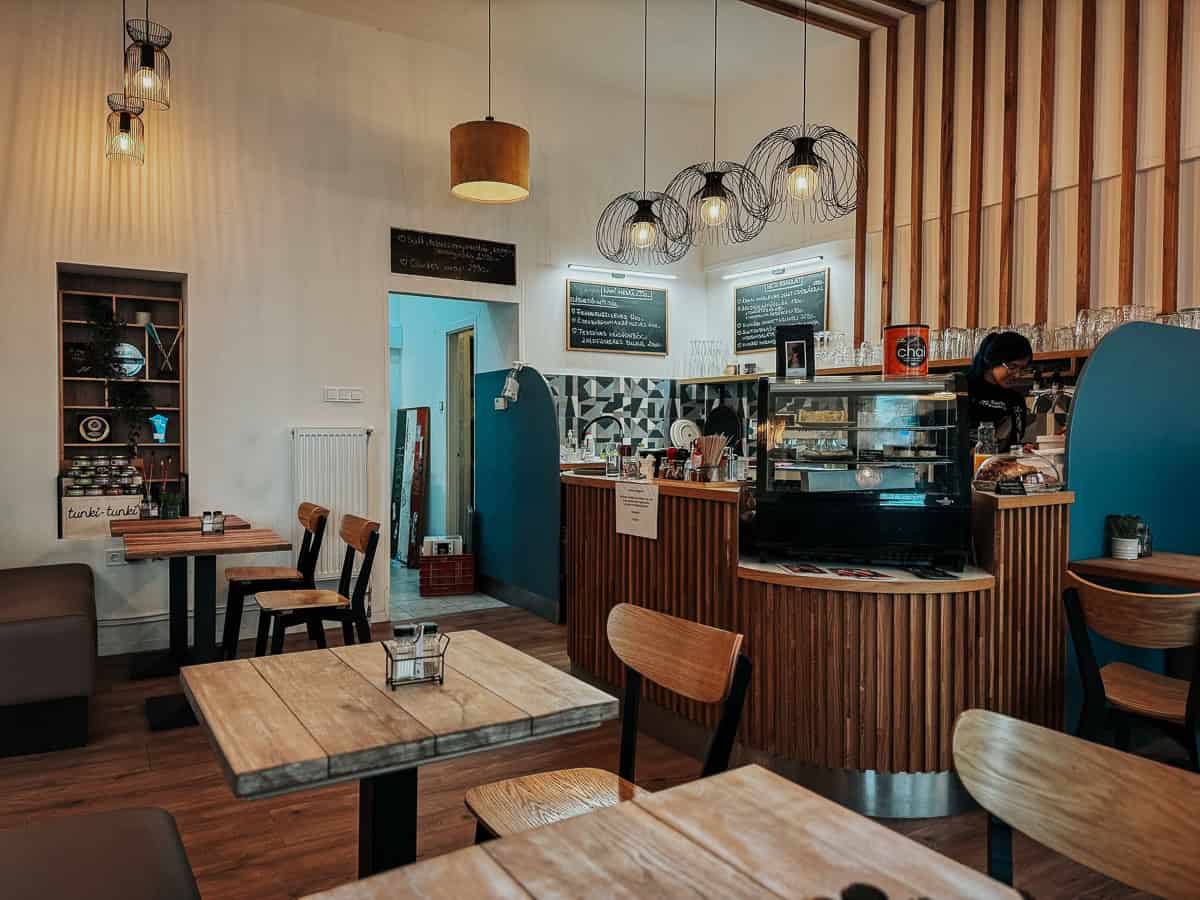 Interior of a cozy café with wooden tables and chairs, warm lighting, and a counter with hanging lamps, where a barista is preparing drinks.