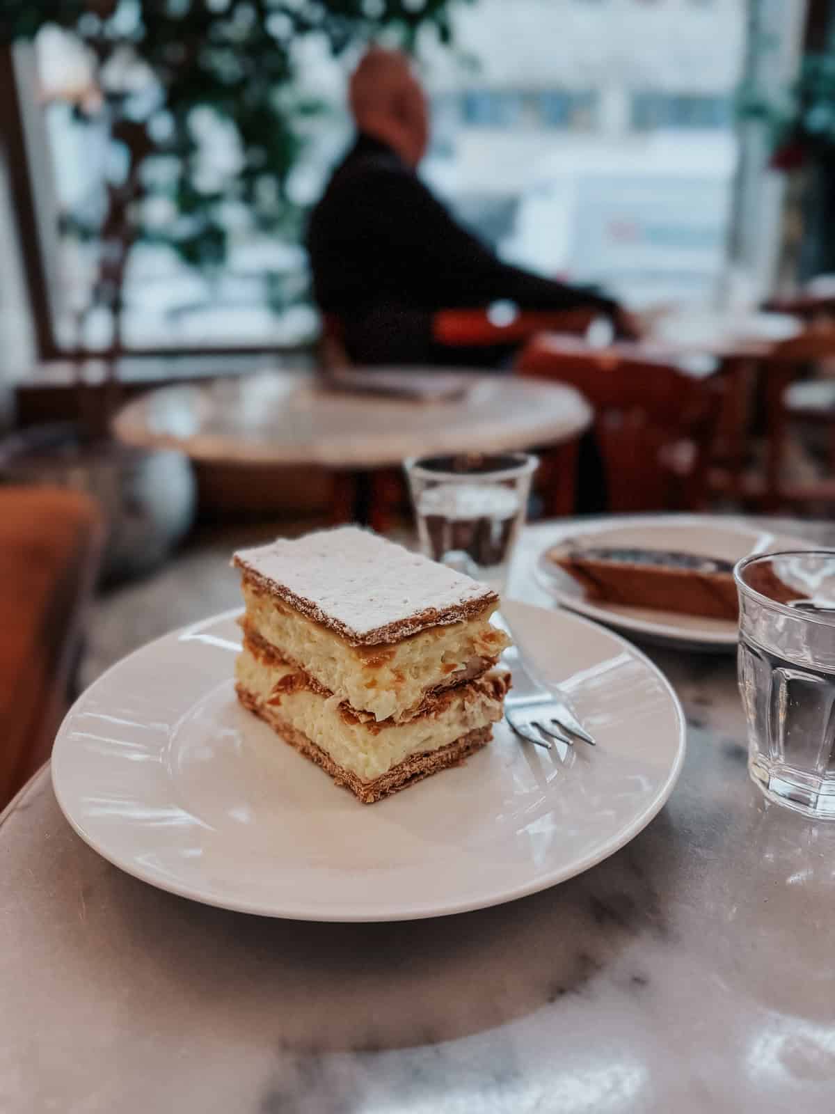 A slice of Hungarian kremes, a layered pastry with cream filling, served on a white plate in a cafe, with a glass of water and a blurred background of a person sitting.