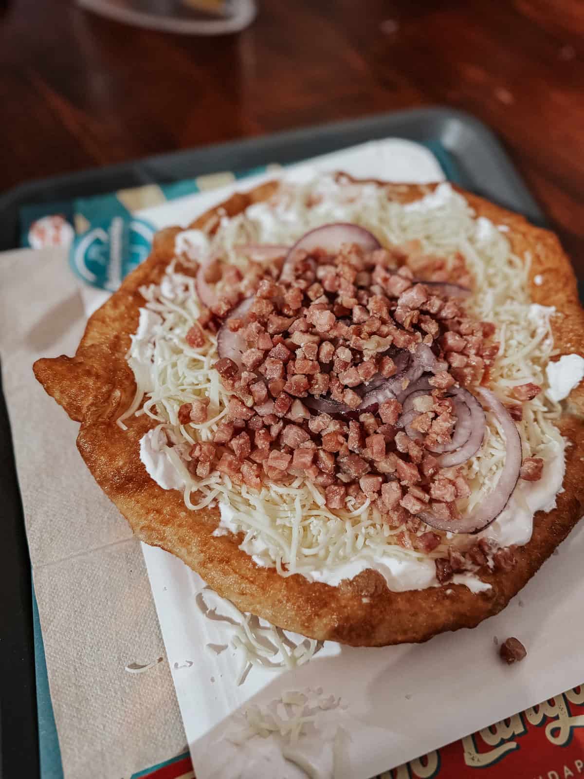 A plate of langos, a Hungarian deep-fried flatbread topped with sour cream, grated cheese, chopped onions, and bacon bits, served on a tray.