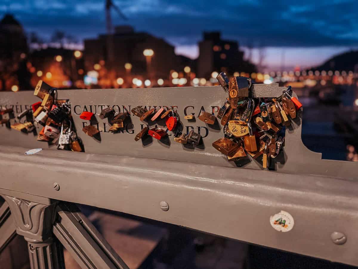 A collection of love locks attached to a bridge railing at night, with the city lights blurred in the background, creating a romantic ambiance.