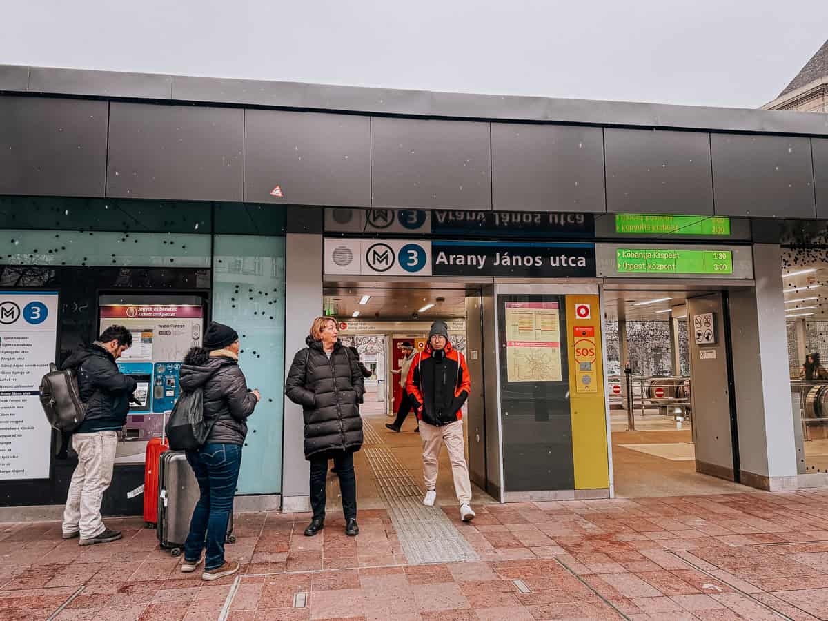 The entrance to the Arany János utca Metro station in Budapest, with passengers standing outside and a ticket machine visible.